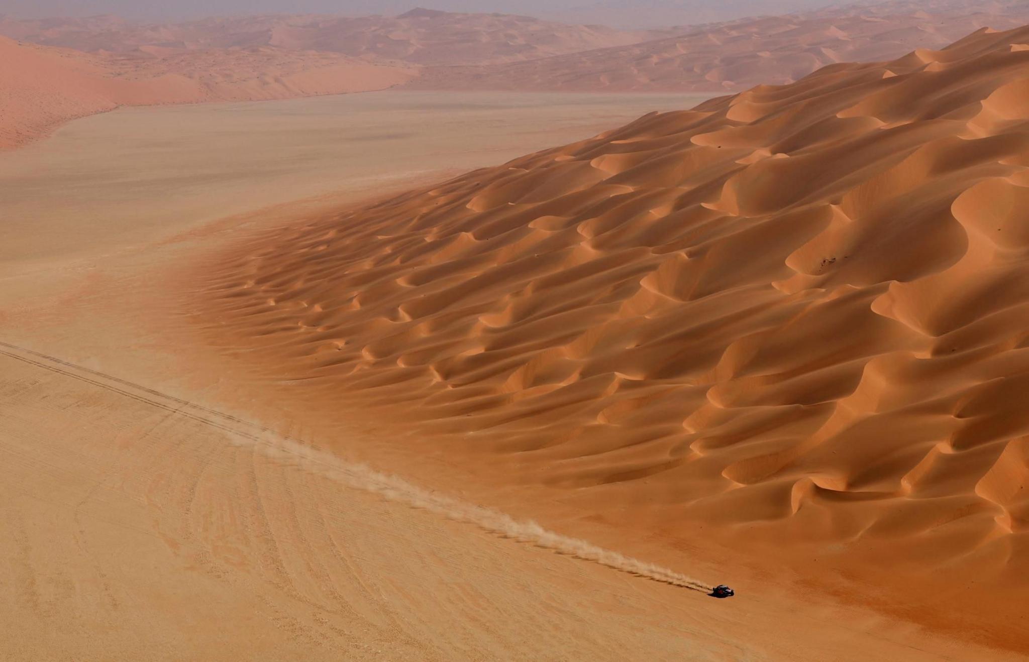 A car drives past large sand dunes, with a trail of dust in its wake