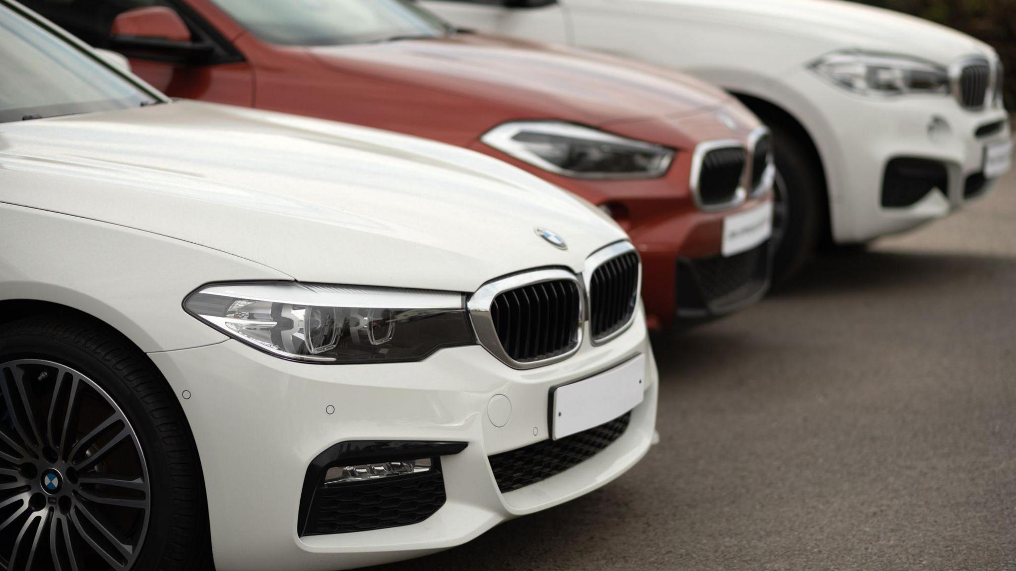A close-up shot shows three BMW cars parked in a diagonal row on a paved surface. The front car is white with a prominent grille and headlights, while a red BMW sits behind it, followed by another white BMW.