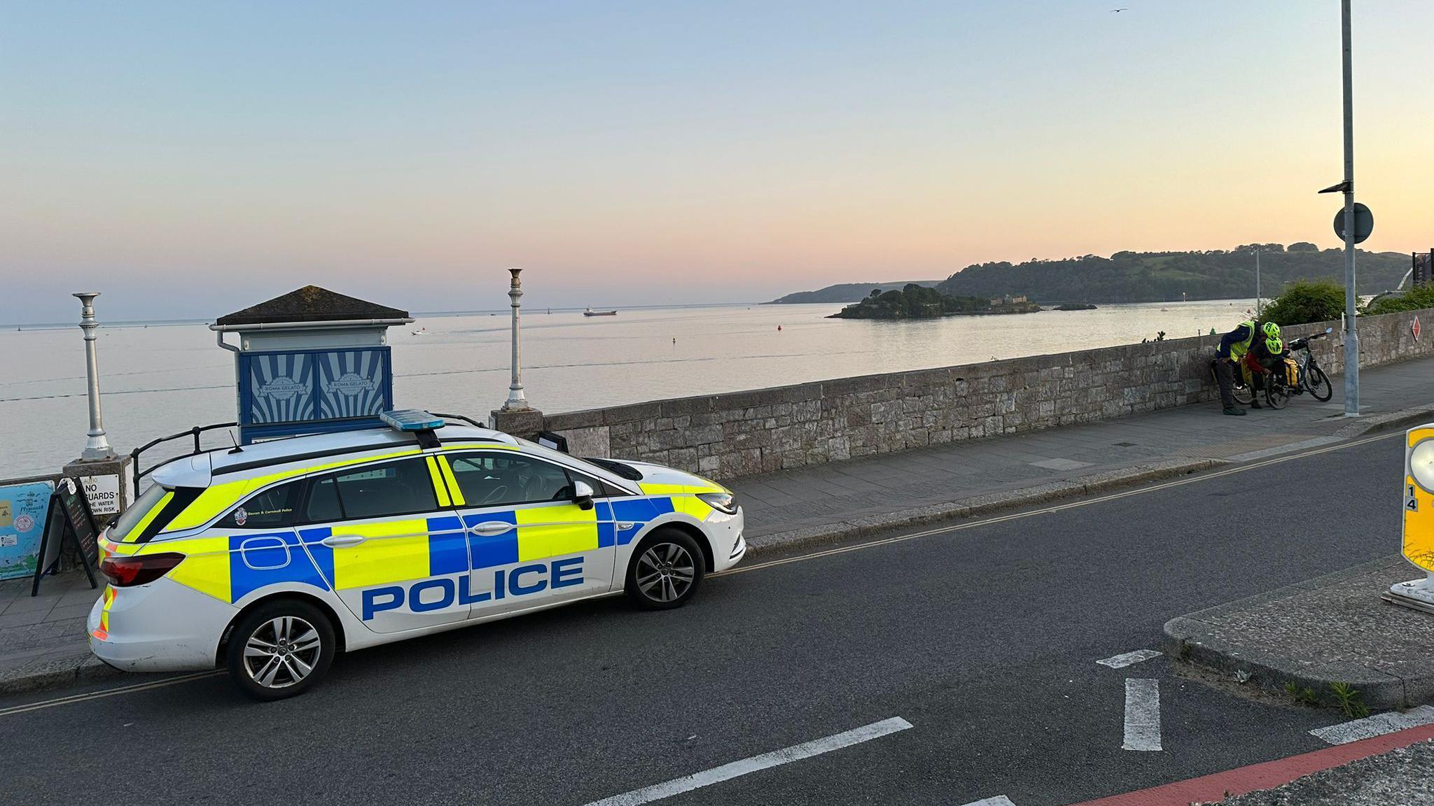 A police car is parked at the side of the road. The road overlooks Plymouth Sound with Drakes Island in the background. The photo has been taken as the sun is setting.