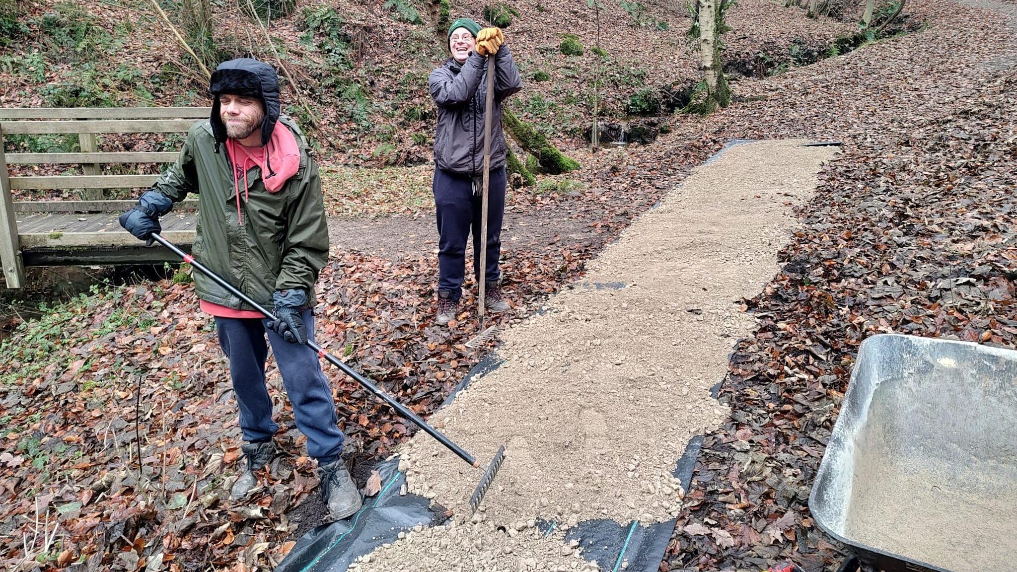 Two people, wearing warm clothing and hats, help to lay a footpath in a woodland. They both have rakes and are spreading gravel over a long rectangular area.