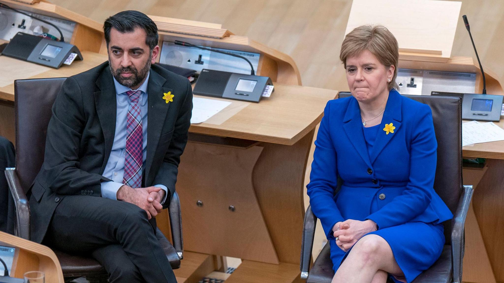 Yousaf, a dark-haired bearded man in a black suit and tartan tie, sits in the Scottish Parliament debating chamber beside Nicola Sturgeon, a brown-haired woman in a royal blue skirt suit. Both are looking off to the side.