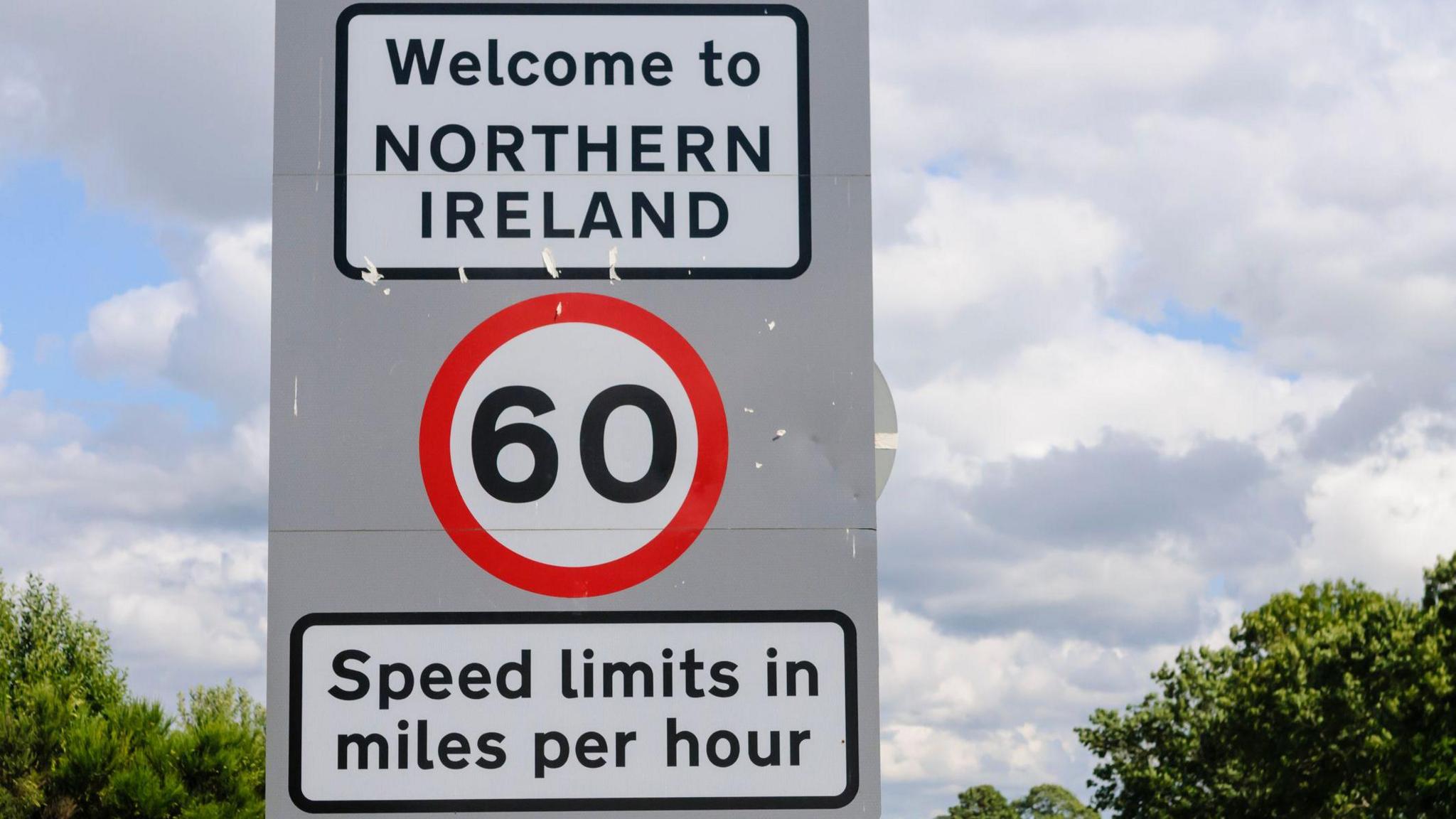 A welcome to Northern Ireland road sign outlining tat there is a 60mph speed limit. There is a cloudy sky in the background. 