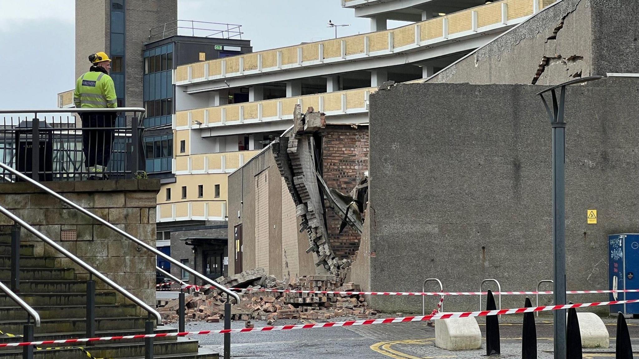 A badly damaged wall with bricks strewn across the ground. An electricity company worker surveys the scene from a balcony at the top of a flight of stairs. Red and white tape has been used to cordon off the scene