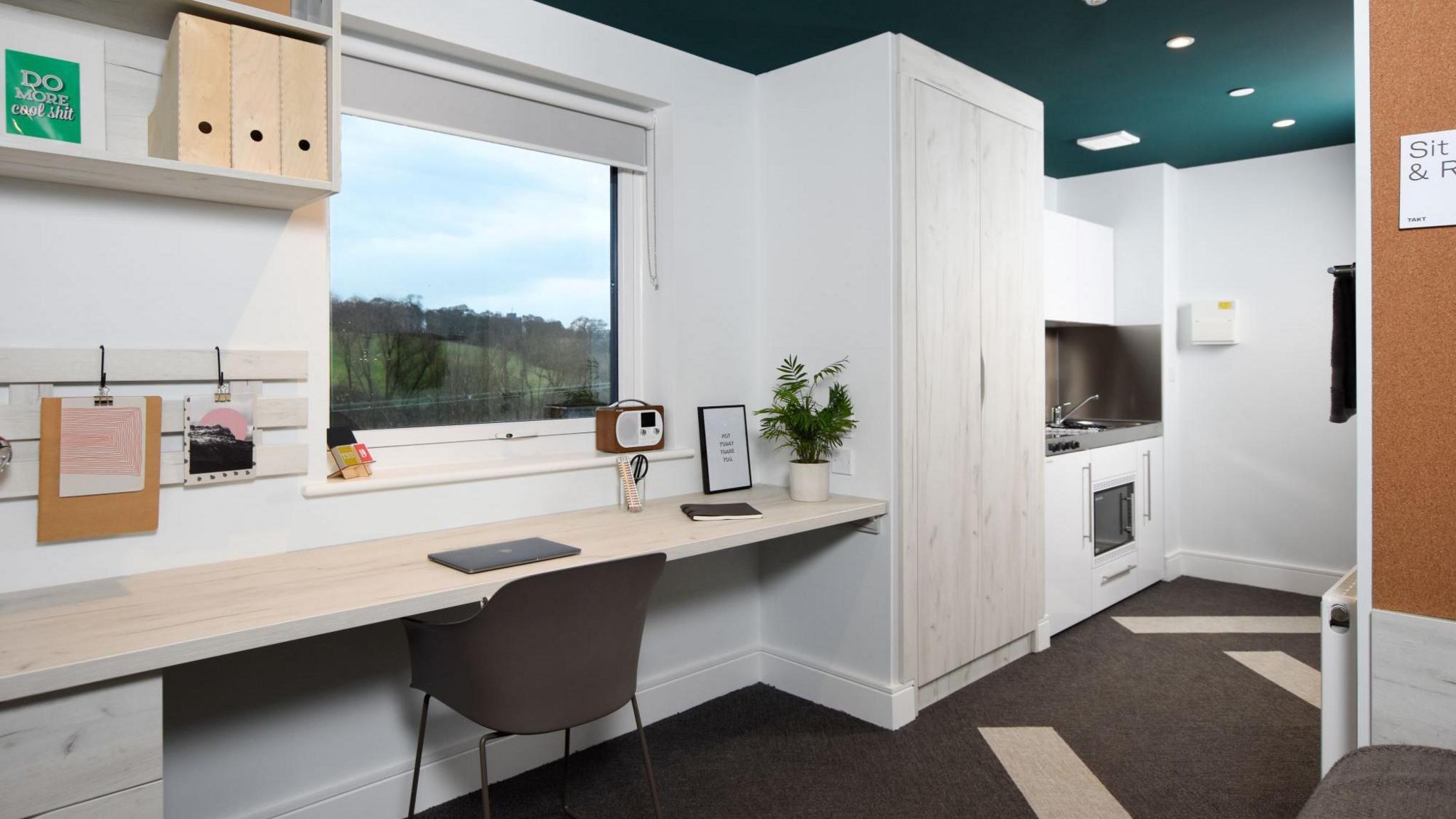 One of the student rooms being used for temporary accommodation. It has white walls and a dark green ceiling. There is a desk, desk chair, wardrobe and small kitchen area. A window offers views of a large field and trees.