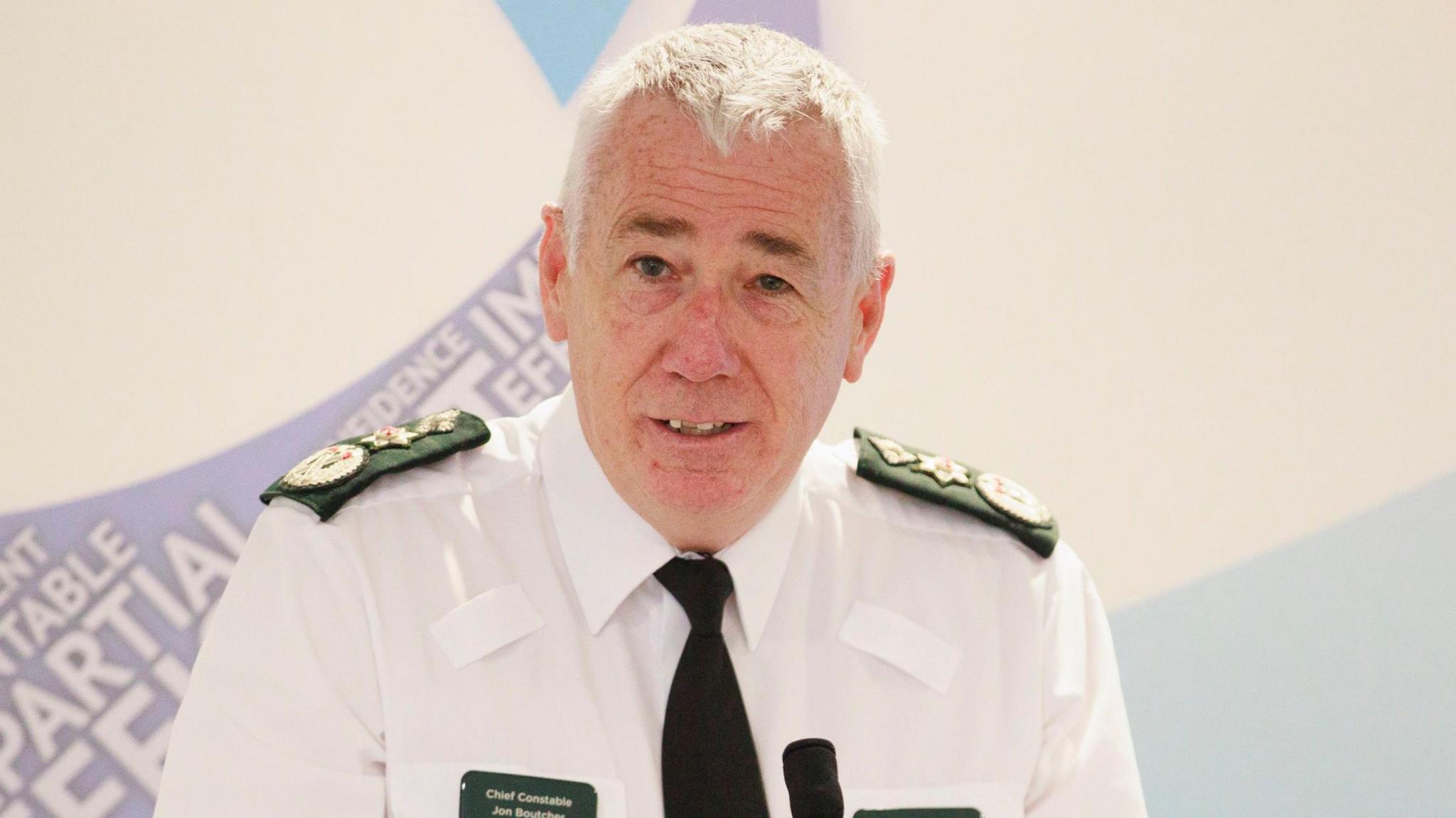 PSNI chief constable Jon Boutcher wearing police uniform - he has white short hair and is looking into the camera as he is speaking at a presser. 