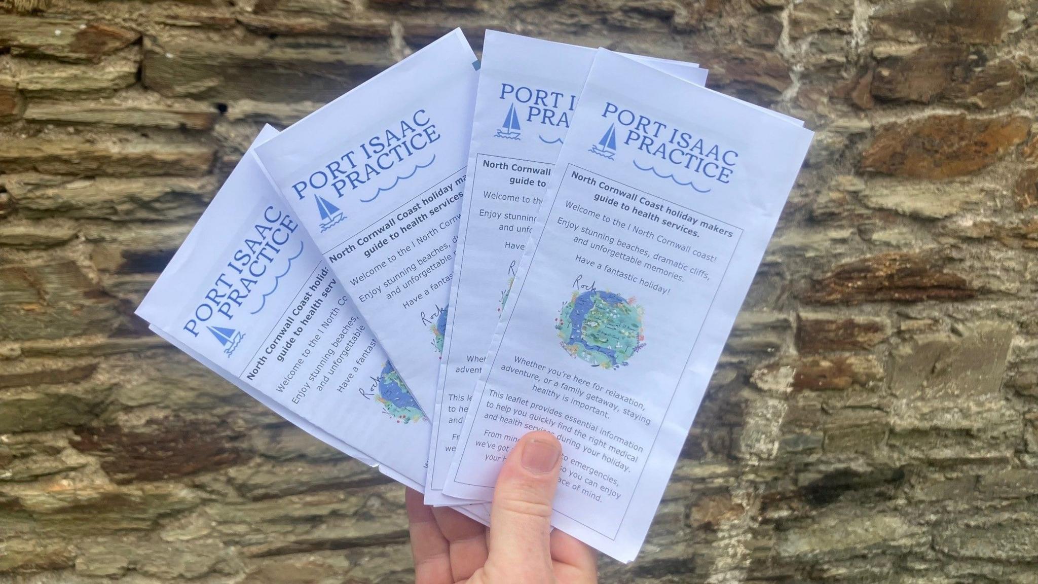 A hand is holding four of the leaflets fanned out in front of a stone wall. On the front is an illustration of the Camel River estuary and the leaflet is headed PORT ISAAC PRACTICE with a sub header reading: North Cornwall Coast holiday makers' guide to health services.