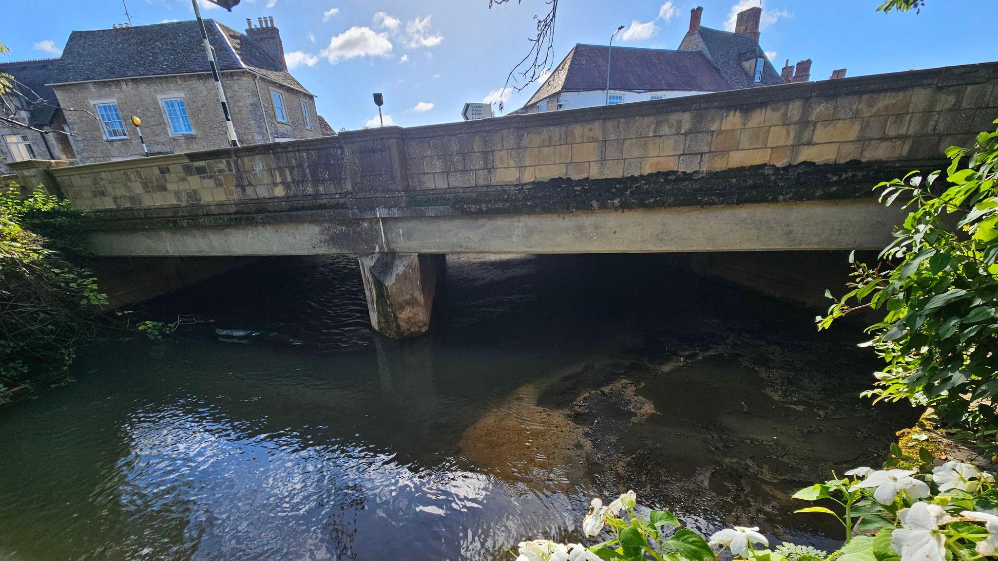 A view of the cleaned-up area under the bridge. The river is flowing. It is a sunny day.