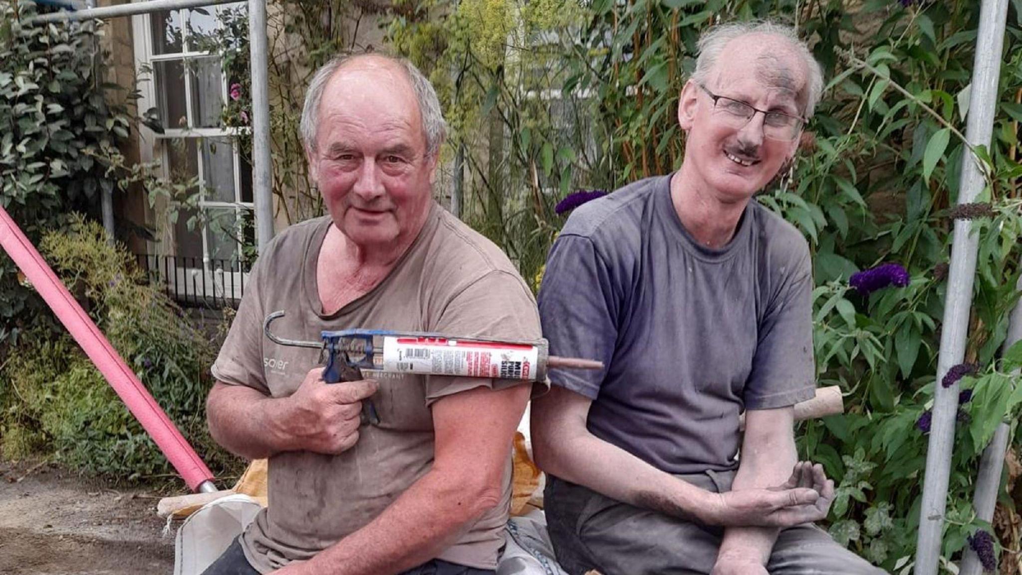 Two workmen in mucky clothes sitting down in front of a house, one is holding a mastic gun