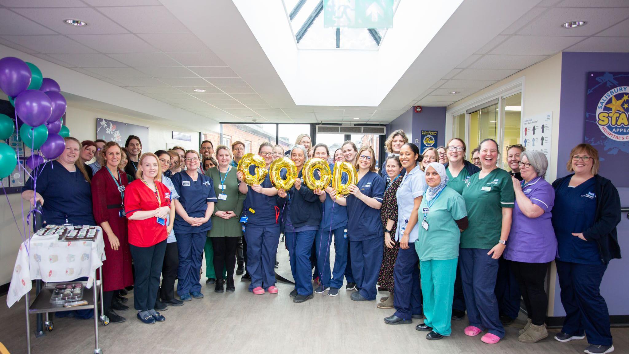 A large group of medical staff holding balloon letters spelling good, all looking at the camera, most wearing medical scrubs.