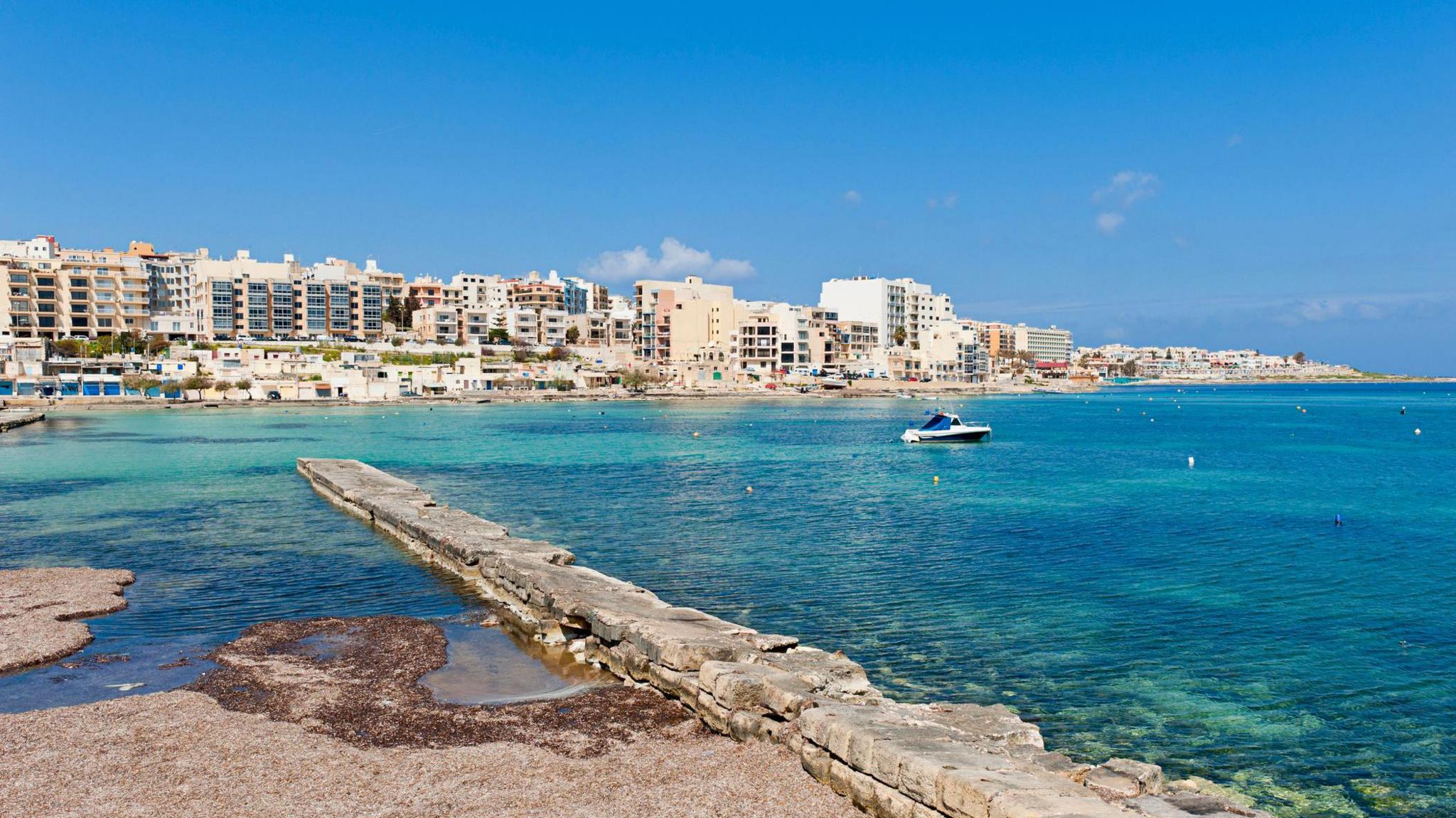 Stone jetty reaching out into turquoise sea with a small boat afloat and a large town sprawling along the coastline in the distance.