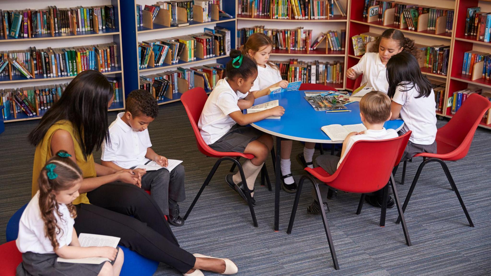 Children and a teacher sit around a table and on soft cushions reading books in a library.