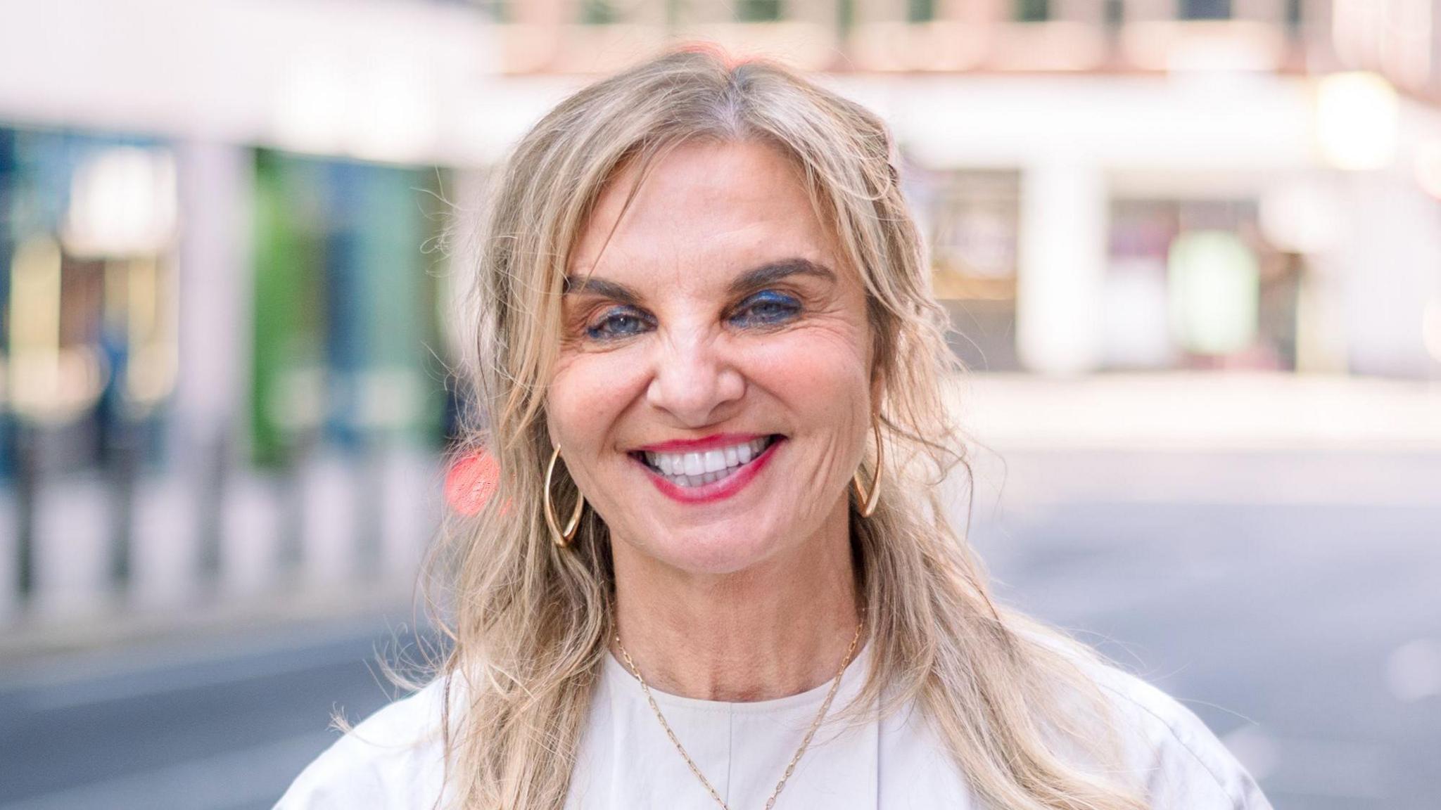 A woman wearing blue eye shadow smiles at the camera. Her shoulder-length hair is worn down and she has gold hoop earrings in. The background is out-of-focus but sunny London street.