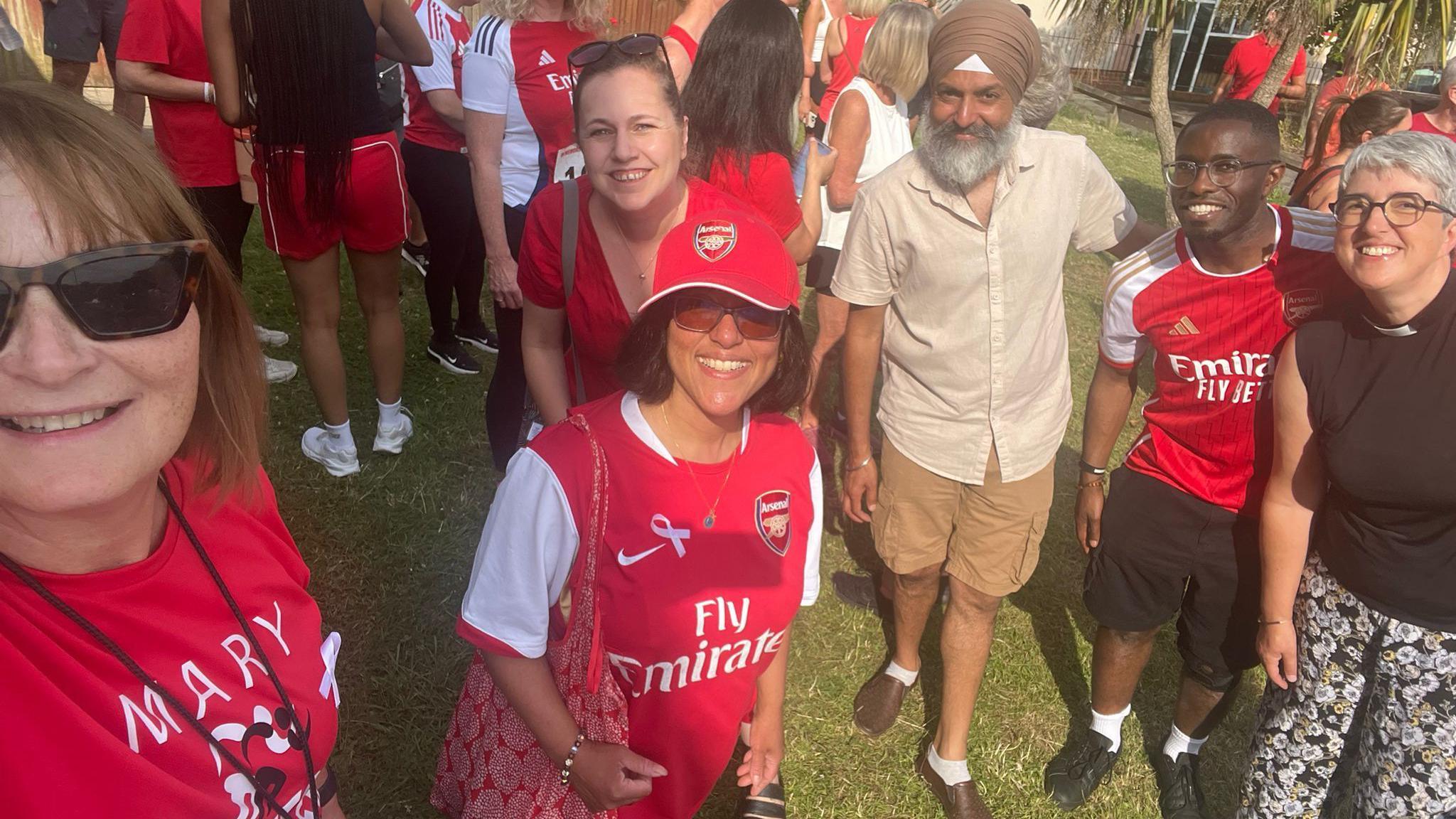 A woman wearing black sunglasses on the left of the photo while she takes a photo. Also in the frame is three woman and two men. 
One woman is wearing a red top and a red sunhat, while another woman behind her is smiling and wearing a red blouse. Next to her is an eldery man wearing a turban, a buttoned up shirt, long shorts and loafers. Next to him is another man wearing a red shirt and black shorts. 
There is a woman at the end of the photo on the right side and she is wearing floral trousers and a black top. They are standing outside on the grass and there are a lot of people outside behind them. 