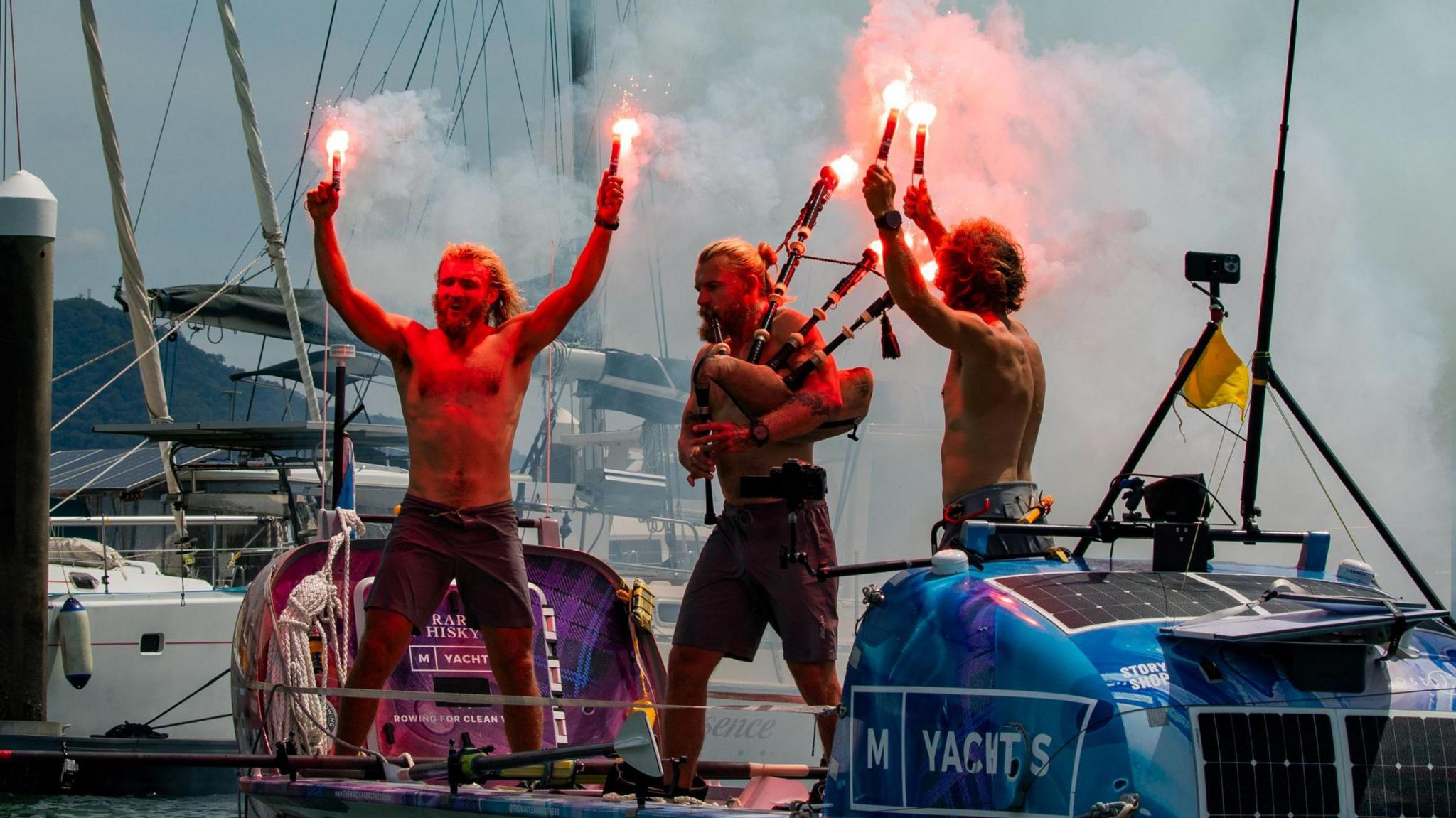 Three happy, bearded, barechested men in a boat wave flares and play bagpipes as they come into harbour on a sunny day.