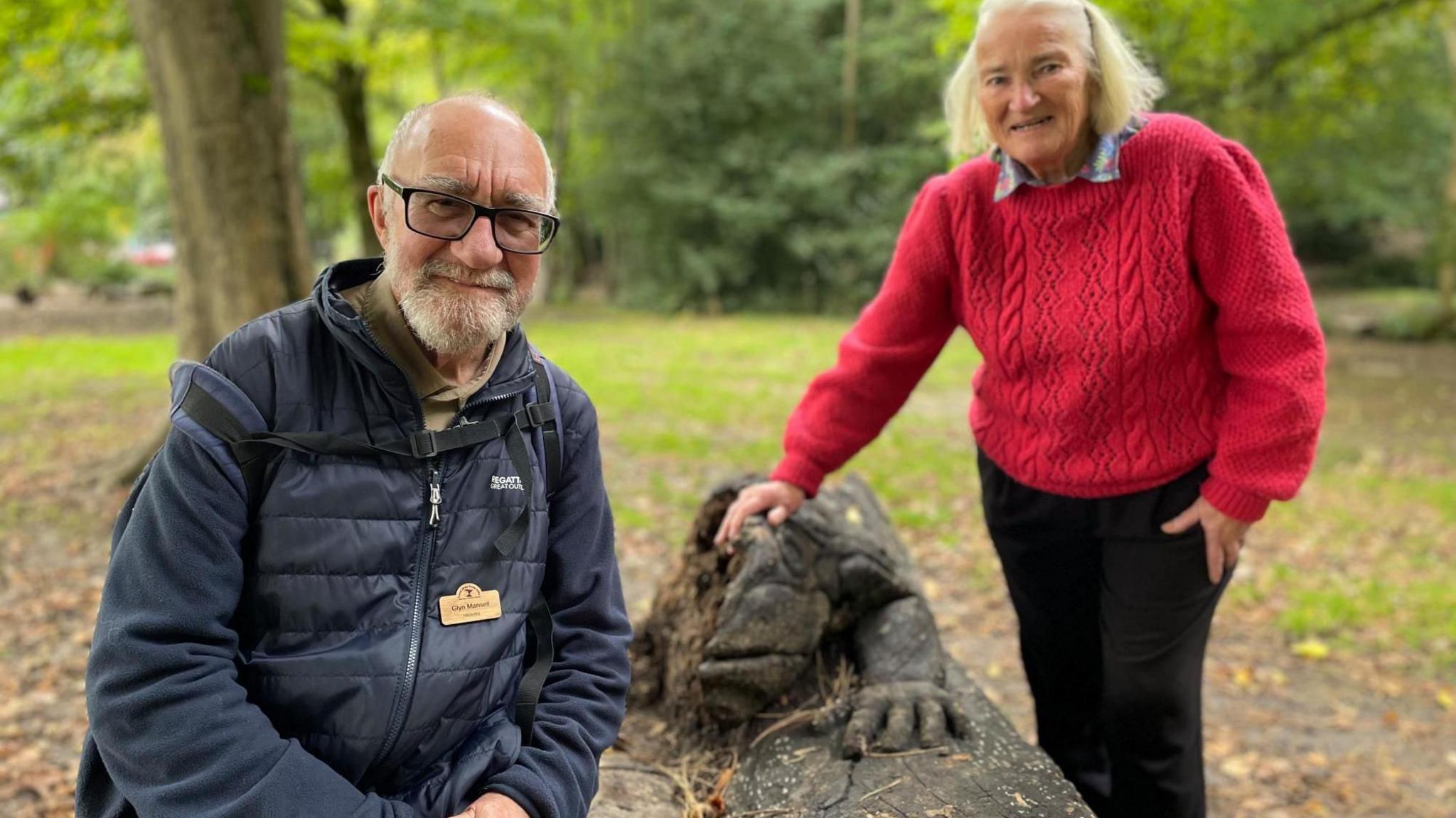 An elderly man and women pose next to a large  wooden toad sculpture in the park