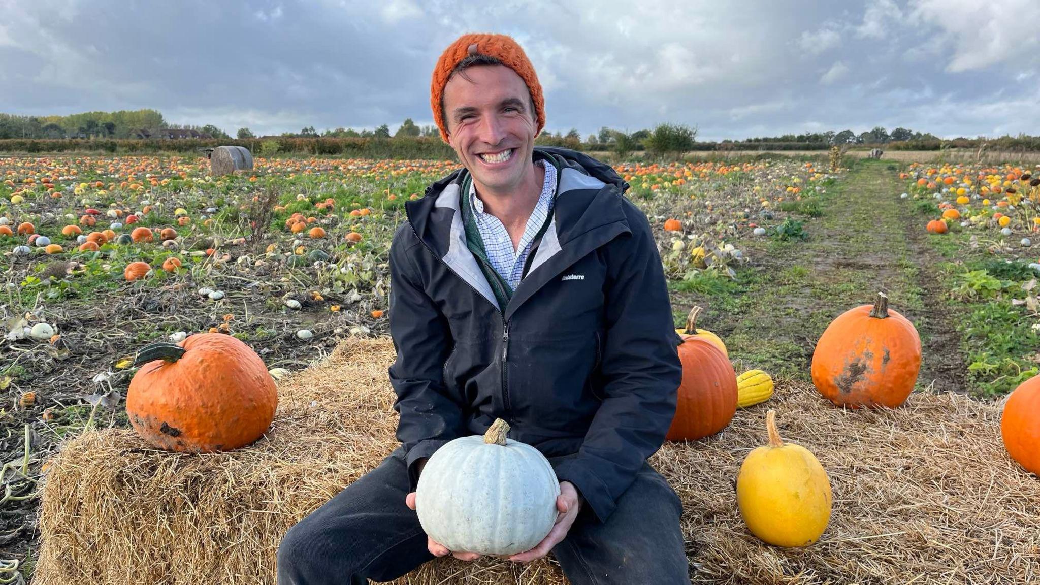 Farmer George Holdstock sat on hay bale in pumpkin field holding grey pumpkin