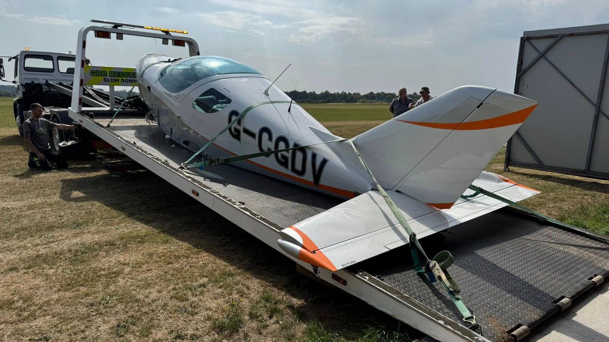 An small aircraft, minus its wings, is sat on the back a low loader. A man is stood to the side operating the lift at the back of the truck, while two others are stood on the other side of the aircraft and truck.