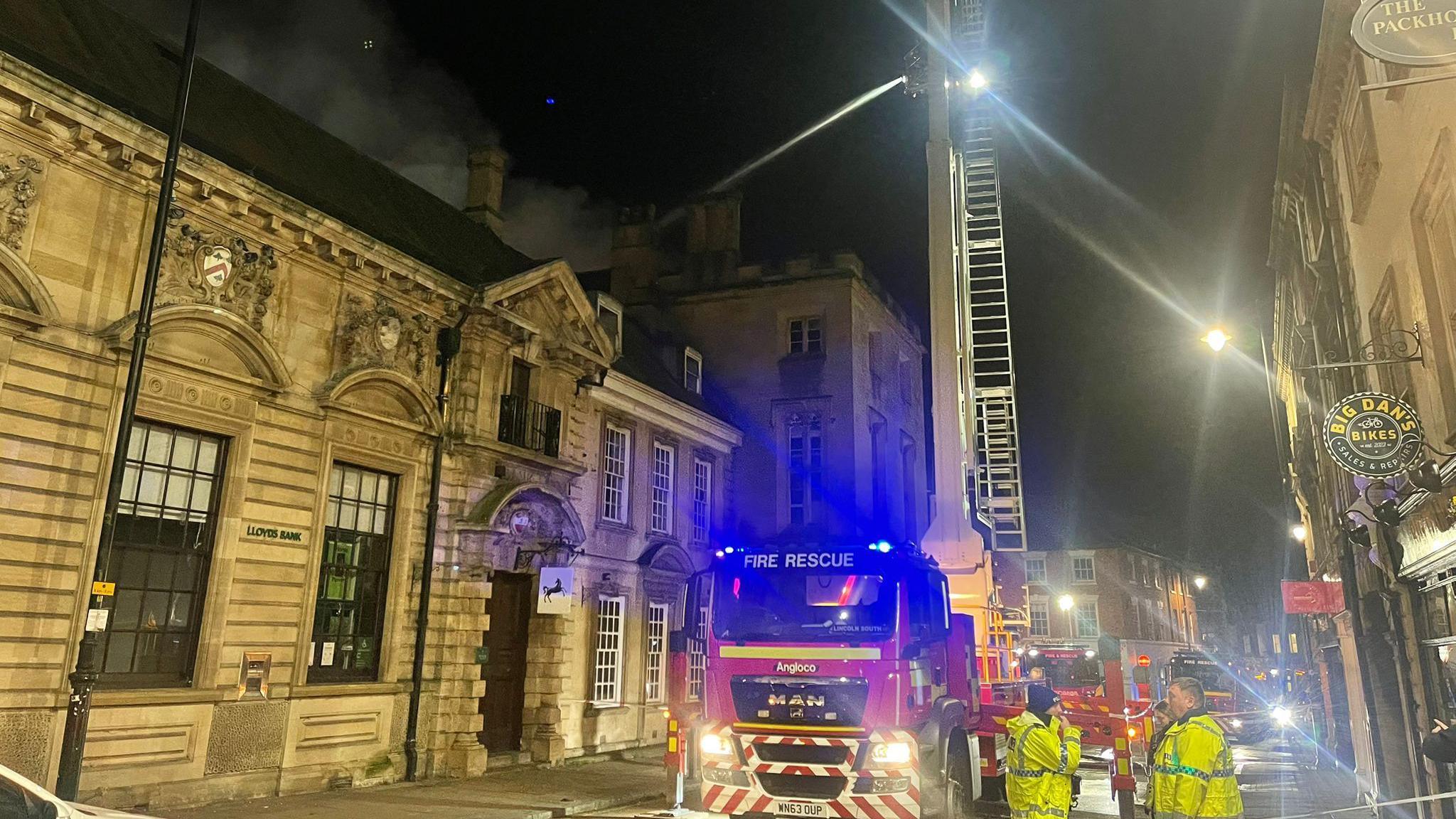 A stone building ablaze at night, with black smoke coming from the roof. A fire engine sits in front of the building. A large fire truck with an extended ladder sprays water. The street is cordoned off with emergency crews present.