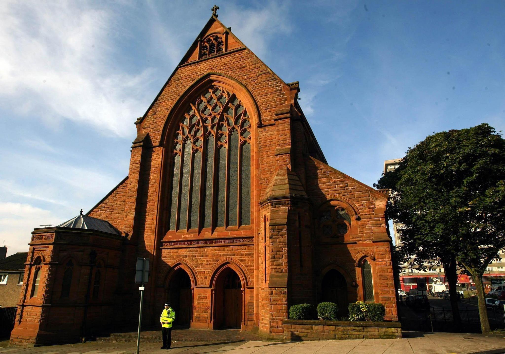 A red sandstone church, in early decorated Gothic style, photographed on a bright sunny day. A police officer stands outside.