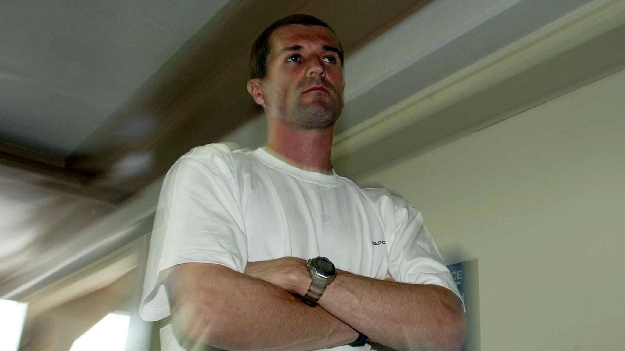 Roy Keane pictured in Saipan at a check-in desk at Saipan Airport. He has short dark hair and is wearing a white t-shirt and a silver watch. He has a unhappy expression, bordering on fed up.