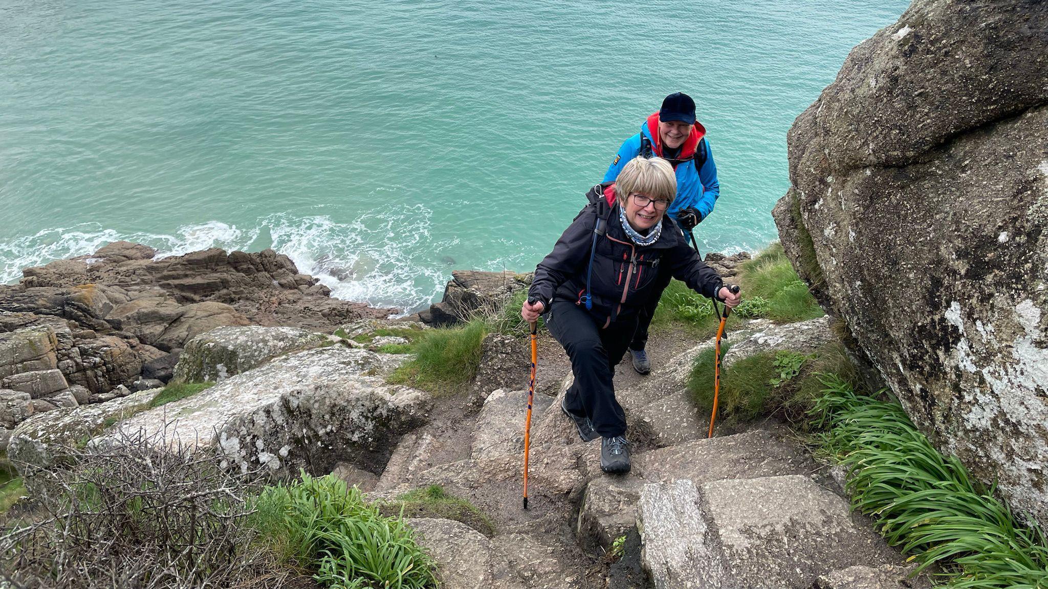 Two Nordic walkers - a man and a woman - walk up a set of large stone steps along the Cornish coast near the Minack Theatre. The sea is behind them. The woman has two orange walking poles and is wearing black trousers and coat. The man behind her has a sky blue coat on and a black baseball cap.