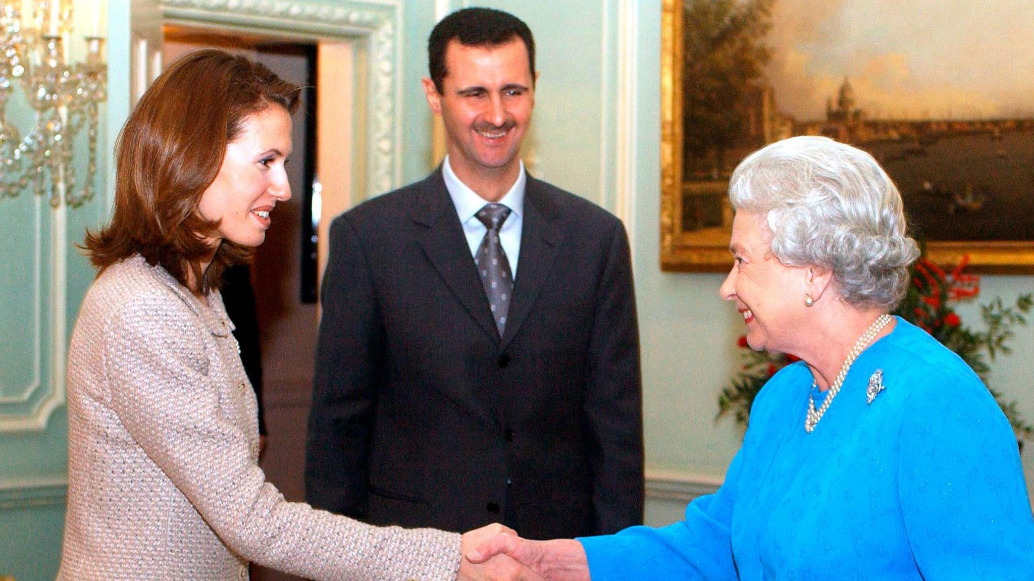 Asma al-Assad, in a pink suit, shakes hands with Queen Elizabeth II, in a light blue suit. Behind their clasped hands stands Bashar al-Assad in a dark suit and tie. All three are smiling. 