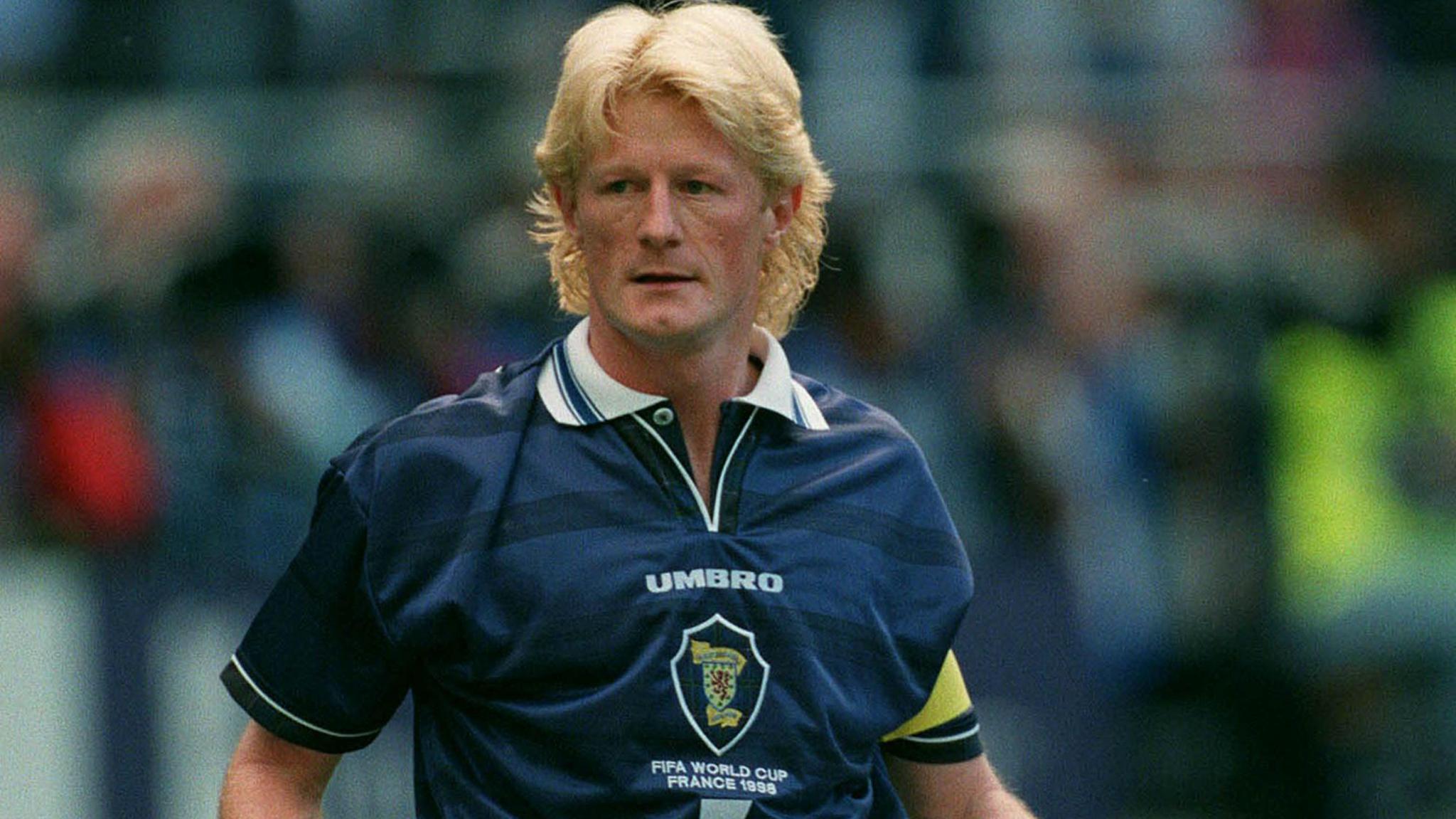 Colin Hendry, with a blonde mullet, wears a dark-blue Scotland short-sleeved football shirt, which has a white collar and the word Umbro and the Scottish badge on it. He is wearing a yellow captain's armband