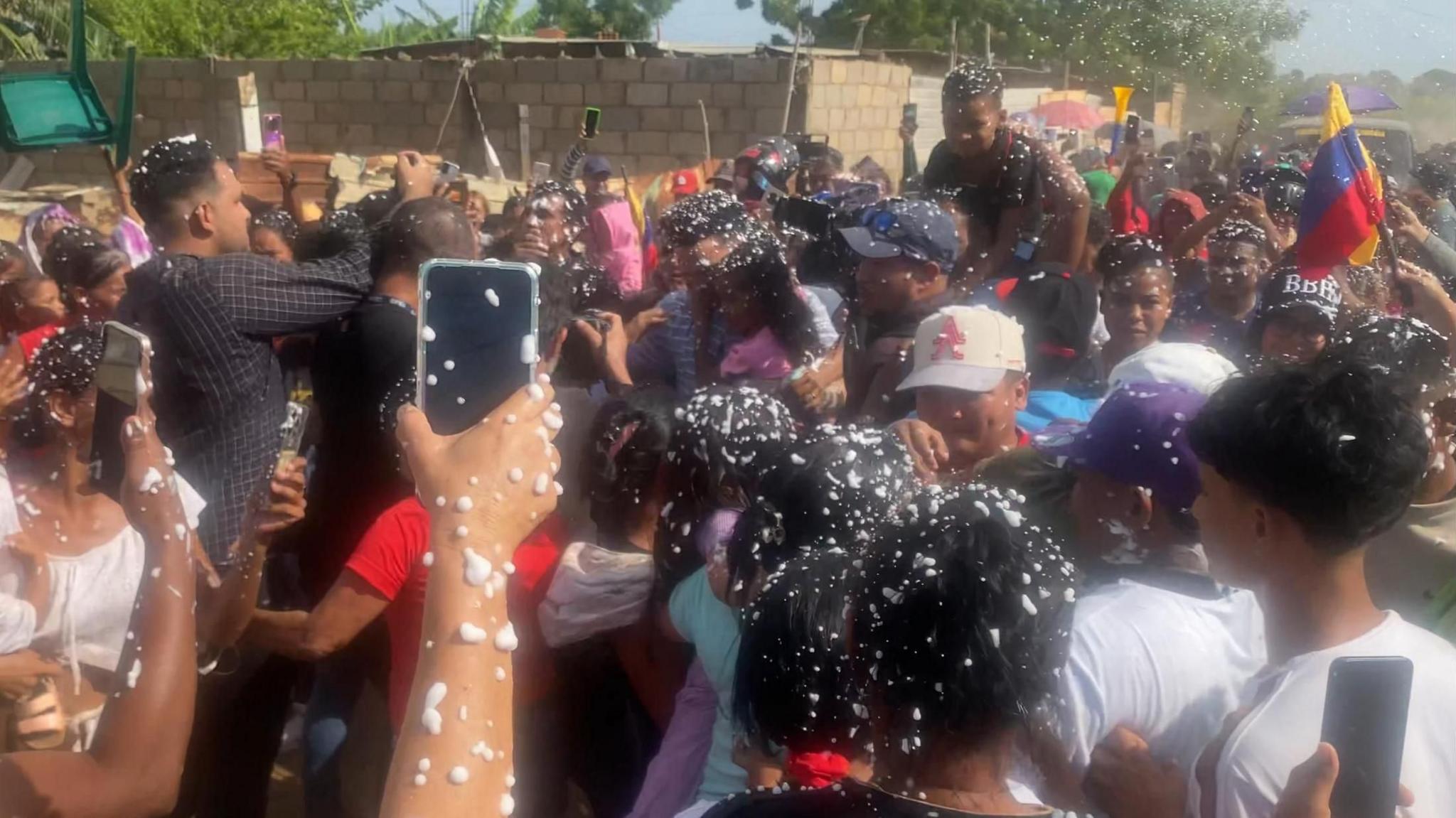 A crowd of people surrounds the men released from detention in El Salvador upon their return to their hometown. Many are covered in foam that someone has sprayed in celebration. Some hold up their mobile phones to record the moment. A child is being held aloft and a Venezuelan flag is waved