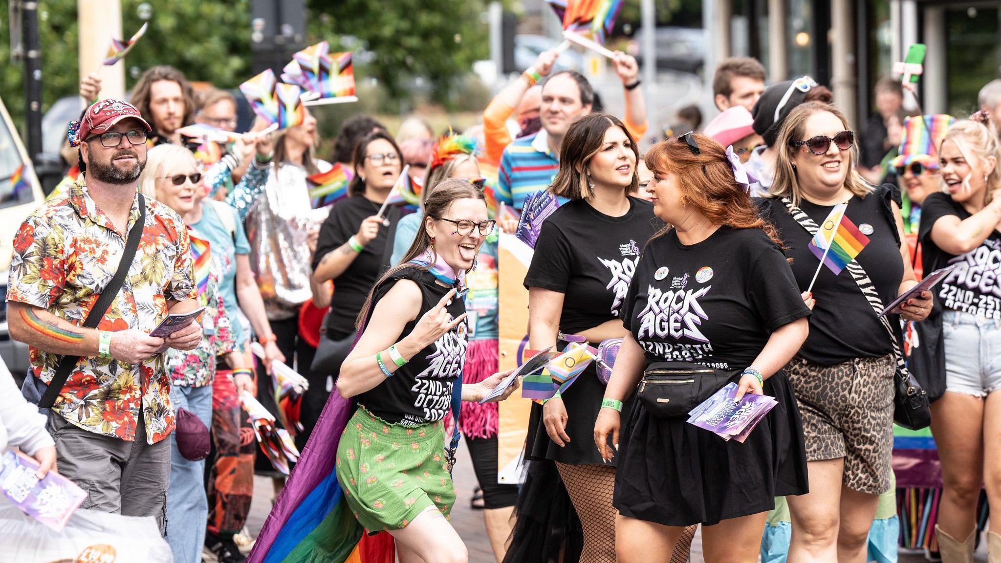 A woman pictured central with a group wearing black Rock of Ages t-shirts does the devil horns hand gesture to a friend, with her tongue sticking out. She is wearing a rainbow cape. Around her are others also wearing colour outfits and waving rainbow flags in a parade.
