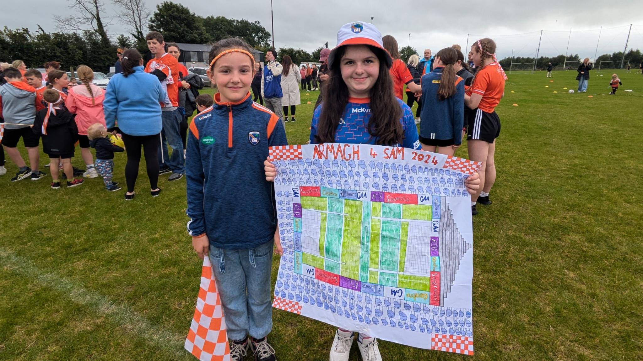 Two girls standing on a GAA pitch, one is holding up a poster that shows a drawing of a birds-eye-view of Croke Park