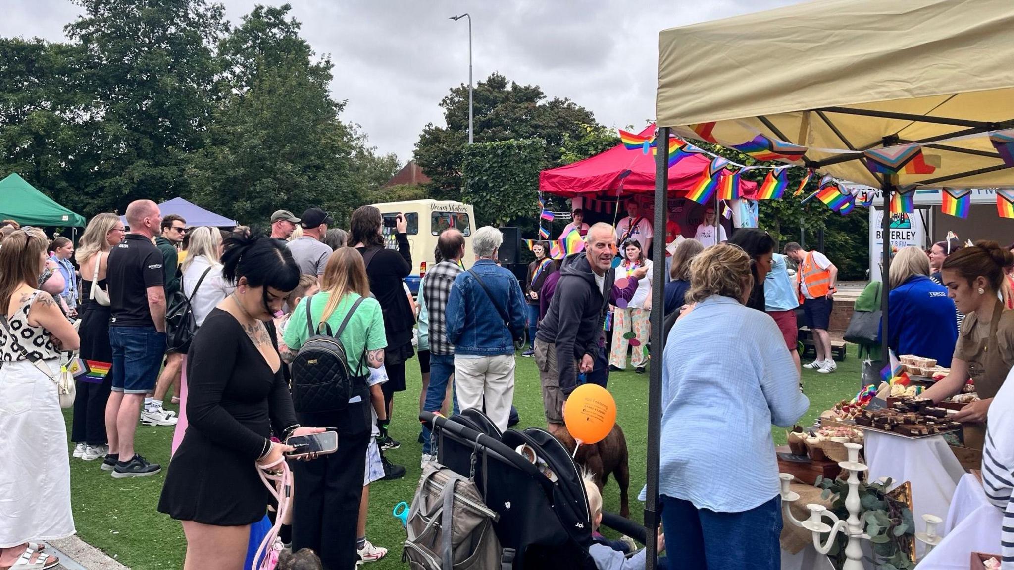 Crowds of people - mostly with their backs to the camera - stood on grass surrounded by market stalls. The sky is overcast.