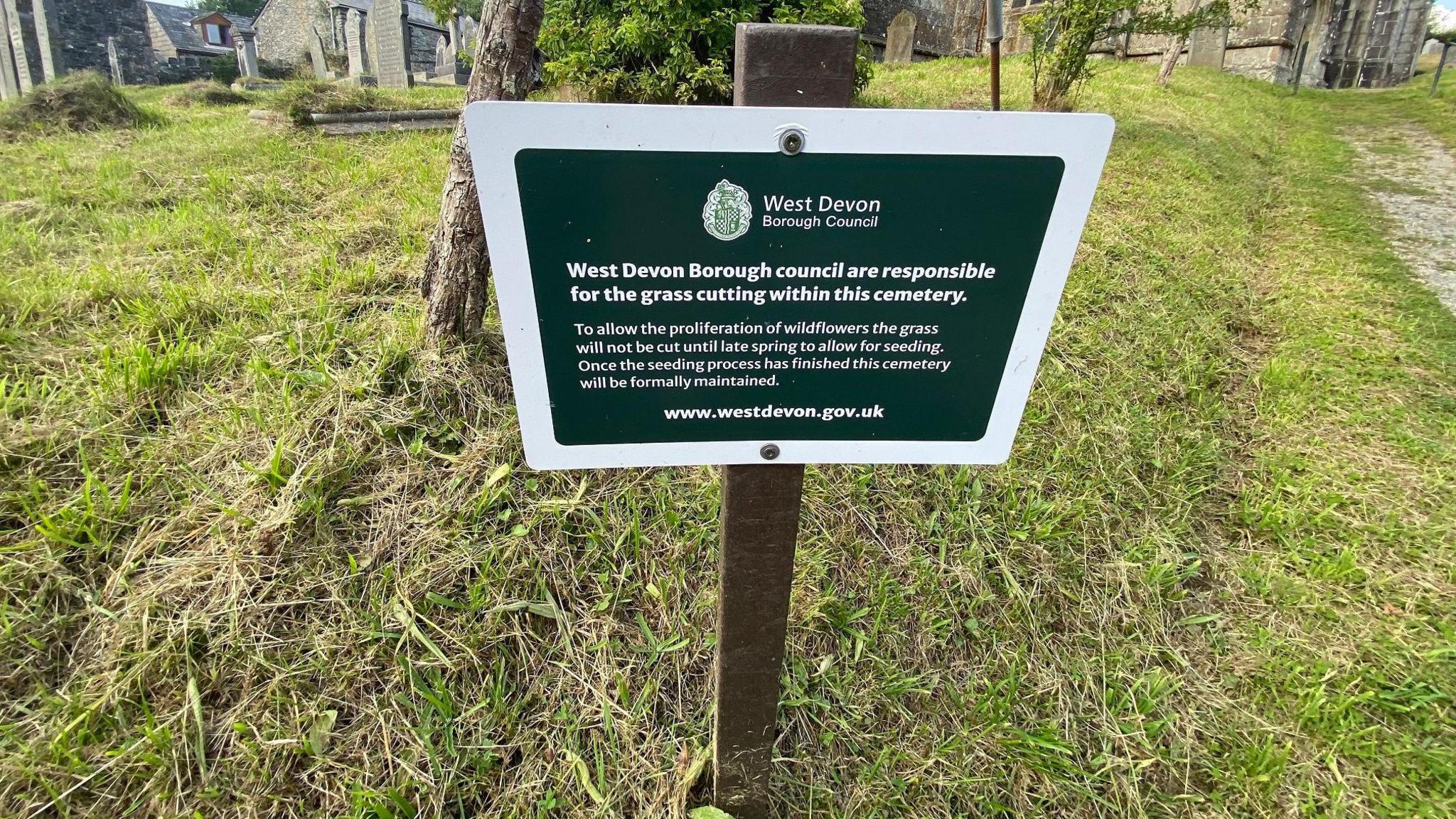 A sign on a post in the closed graveyard at the St Mary parish church. It is headed West Devon Borough Council and the first line says: West Devon Borough Council are responsible for the grass cutting within this cemetery. It goes on to say that to allow the proliferation of wildflowers grass cutting will be limited. The website www.westdevon.gov.uk is at the bottom of the sign.