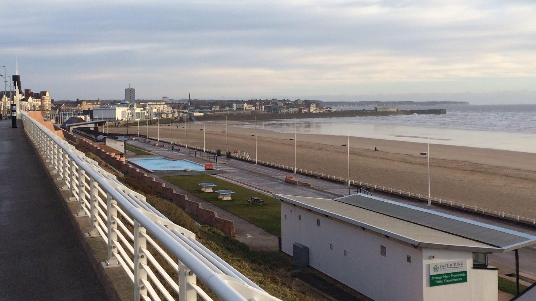 A view of Bridlington's South beach, with a promenade and a large sandy beach, with the sea to the right. In the background, large buildings and a harbour, with the coast sweeping around to a headland.