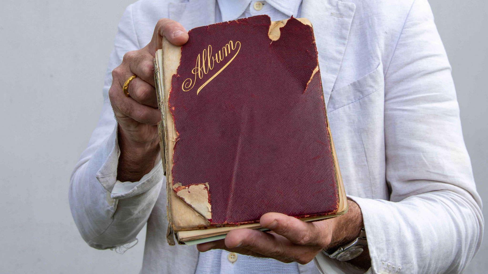 An old book with a worn, red cover and gold writing saying 'Album' being held by a man wearing a white overcoat.