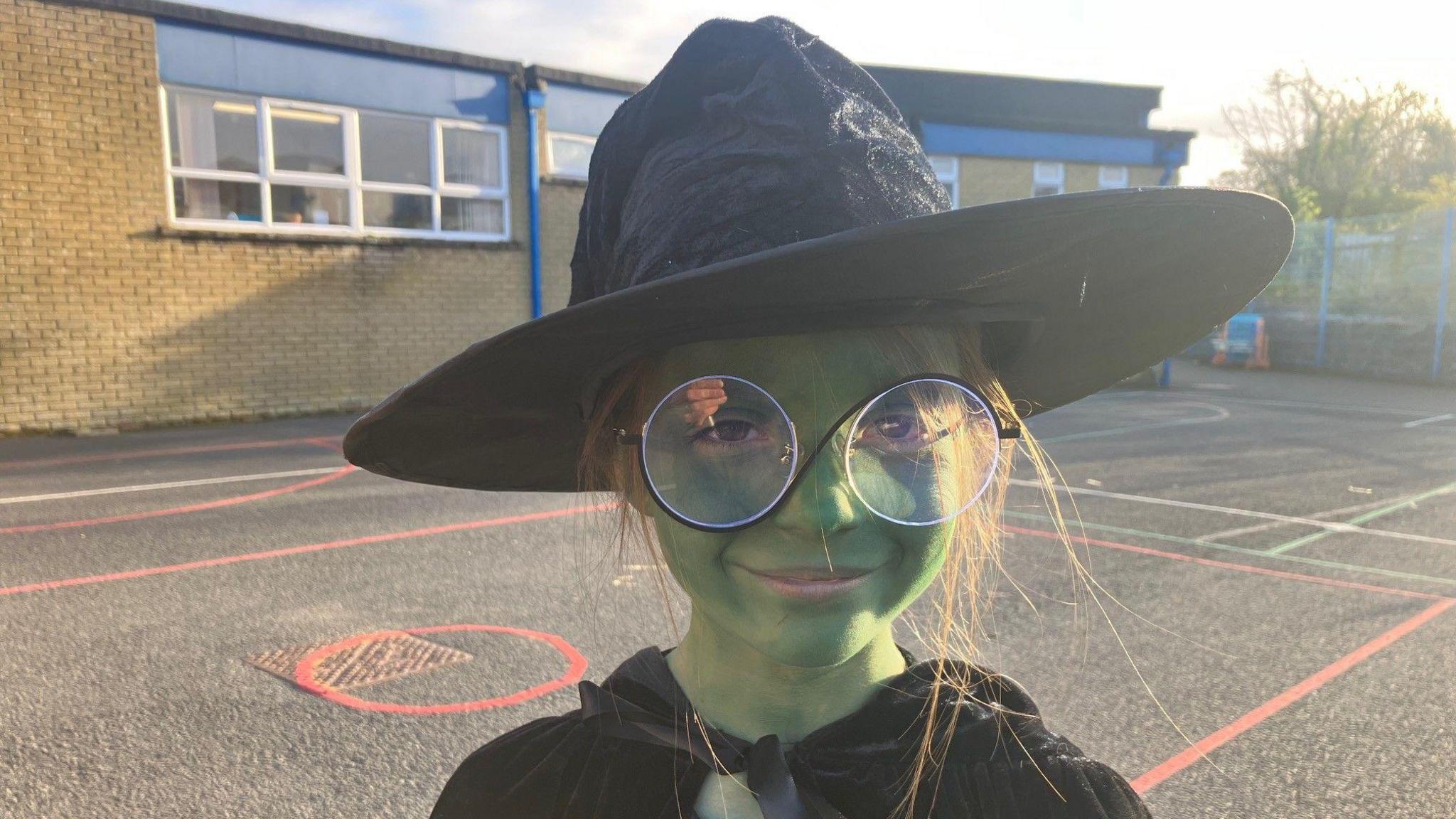 Ella Rose is dressed up like the Elphaba from Wicked. Her face is painted green and she's wearing a witch hat and a black cape. She's wearing large circle-shaped glasses. She's outside in a school playground.