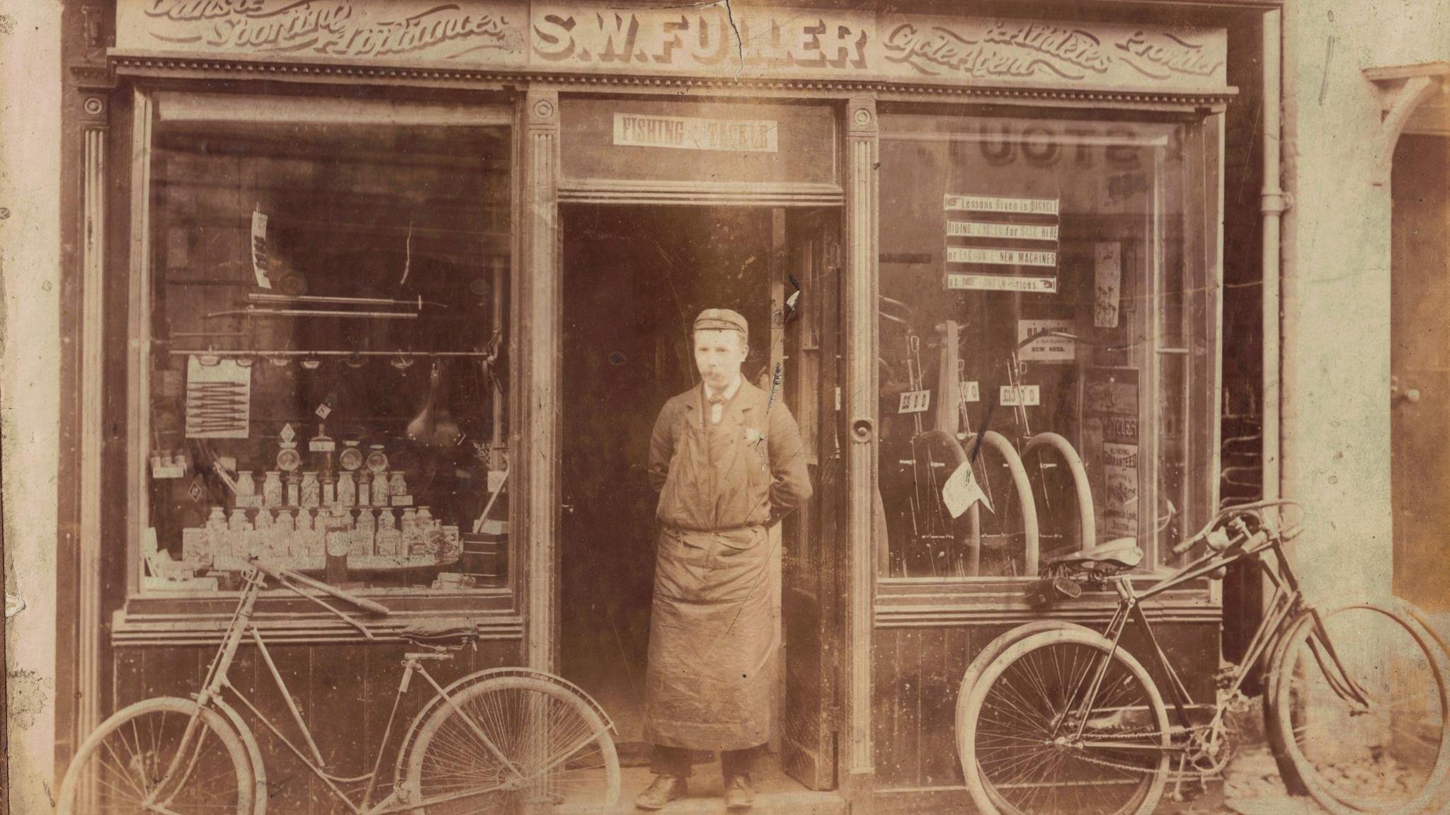 Sepia photograph of a man in a suit and lare apron standing in the doorway of S W Fuller shop. 