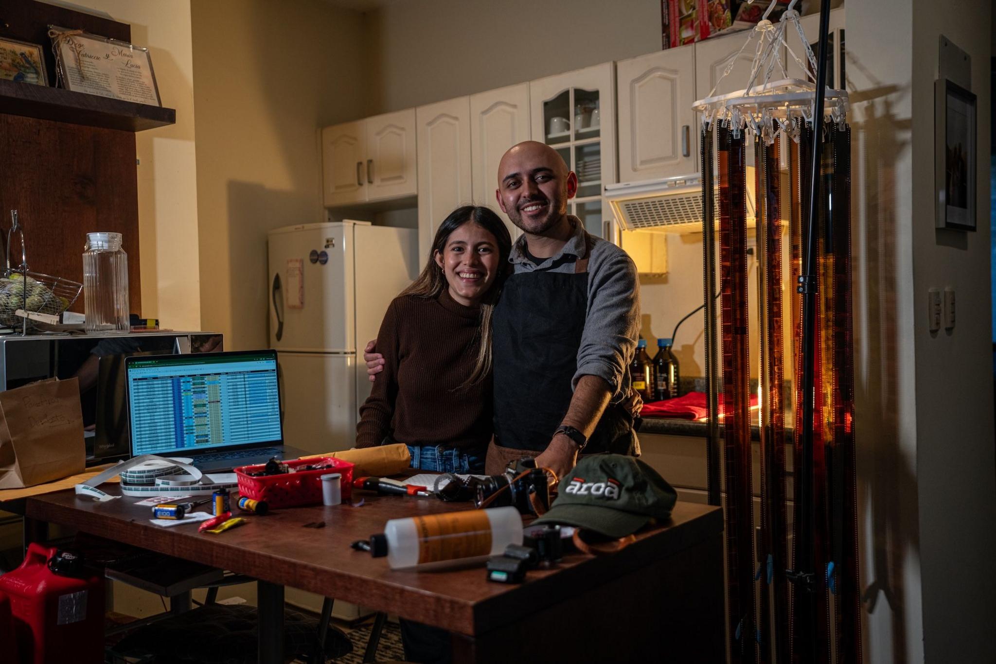 Fabriccio Díaz and Lucía Ramírez embrace in their kitchen where they develop all the film for the Arca Film Lab. On the desk in front of them, bottles of chemicals can be seen and rolls of film hang drying. 