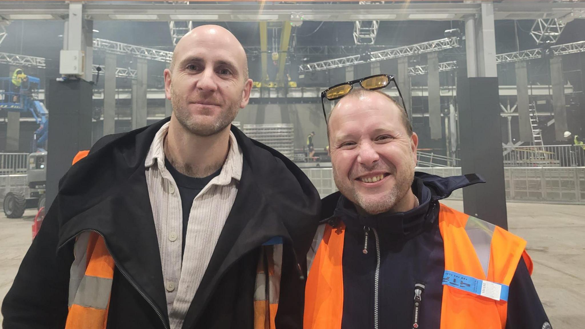 Directors of AMAAD, William Harold and Paul Sobierajski. They are standing in the empty Prospect Building while it is being set up behind them. They are wearing orange high vis and dark clothes.