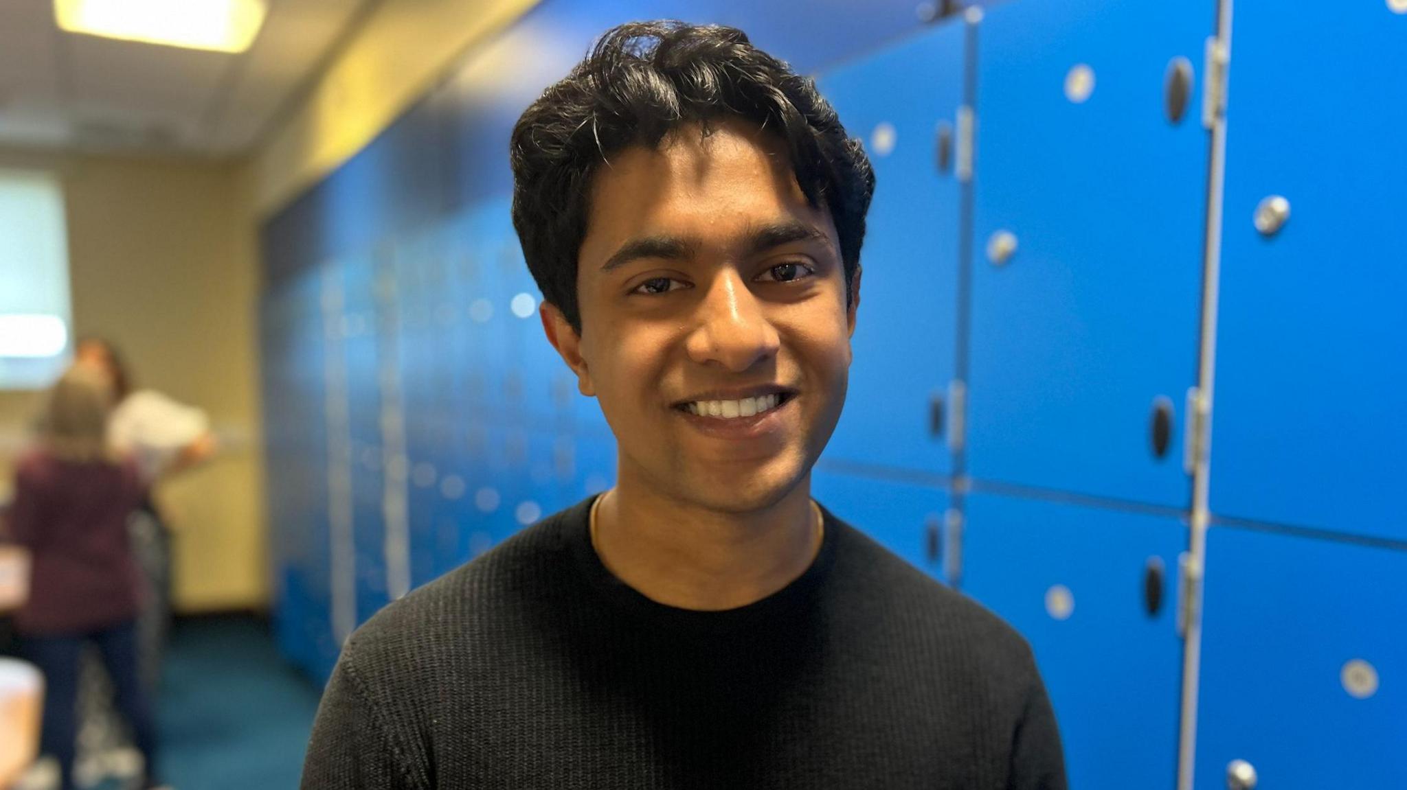 Praveen Ballakrishnan smiles at the camera, he is wearing a black top and is standing next to blue lockers.