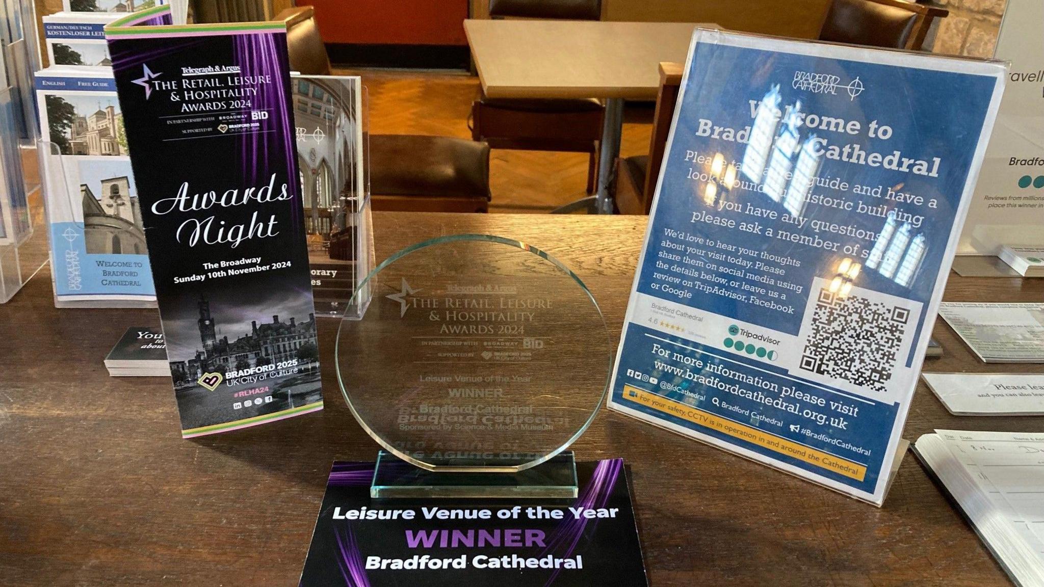 A circle-shaped glass trophy on a brown, wooden table with an awards night  booklet on the left and a blue and white laminated Welcome to Bradford Cathedral poster. 