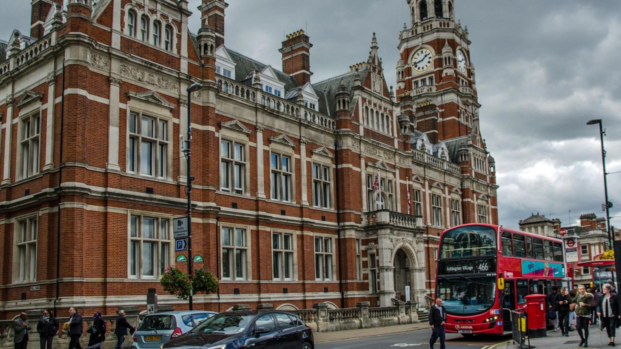 Pedestrians and traffic passing the red brick Town Hall headquarters of Croydon City Council, there is a clock face on one of the building's columns.