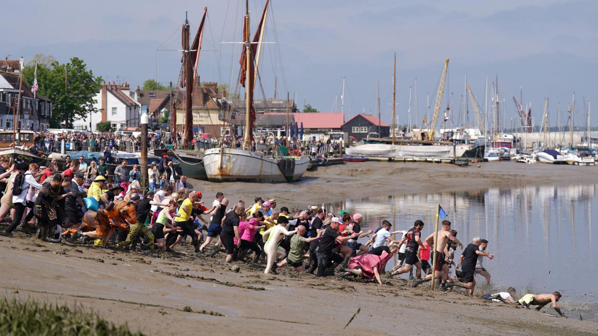 Hundreds of people, many in fancy dress, are running through mud into Hythe Quay in Maldon. There are boats in the background and historic buildings.