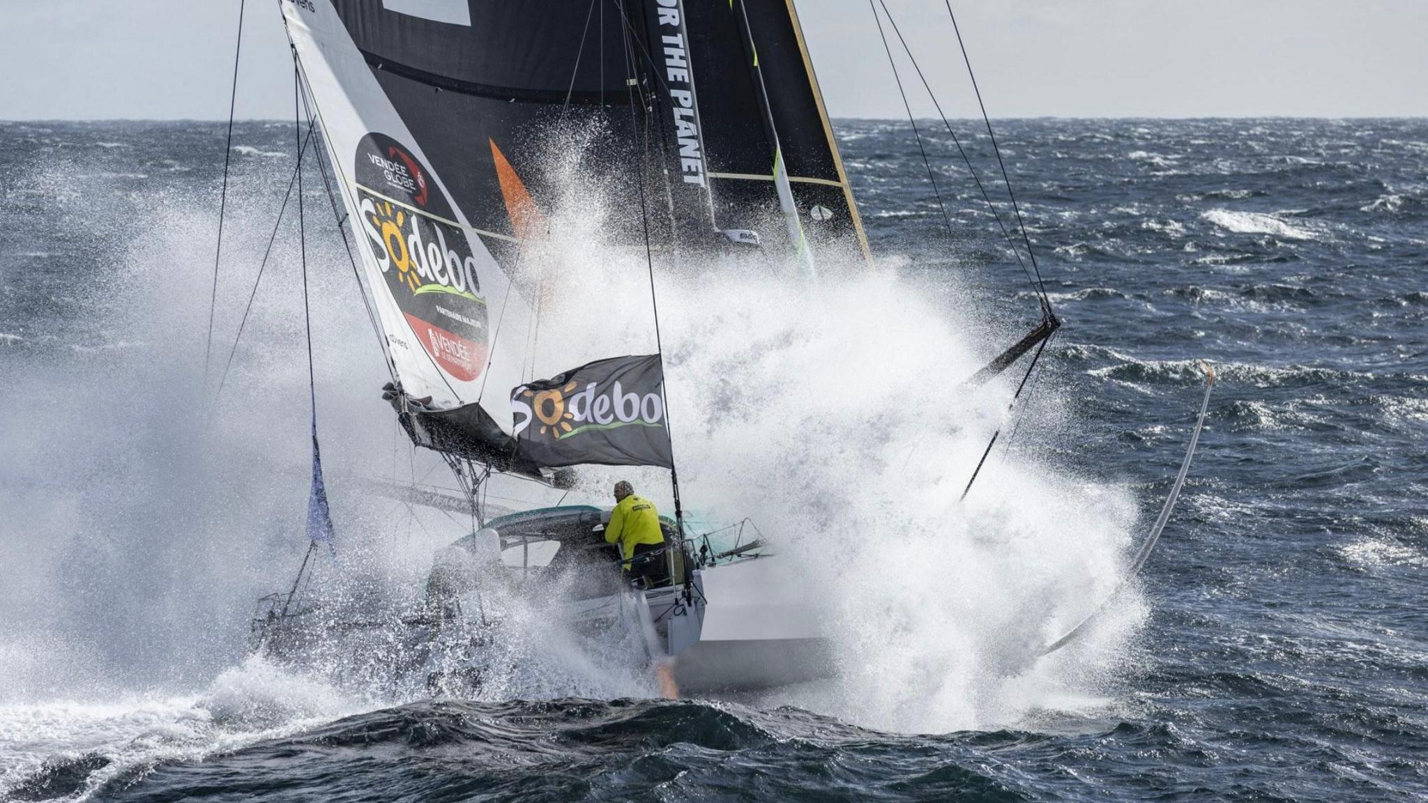 A sailing boat in rough seas with the word Sodebo on a sail and a sailor in a yellow top in the cockpit.