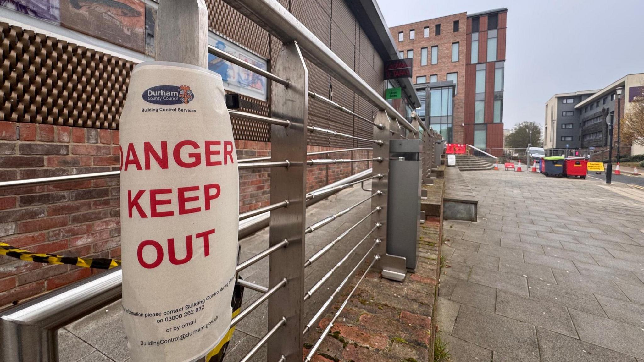 A sign which reads 'DANGER KEEP OUT' in red letters, with the Durham County Council logo at the top, attached with black and yellow tape to railings near Durham Passport Office. In the background, cones and temporary metal fences are visible on the path adjacent to the office. 