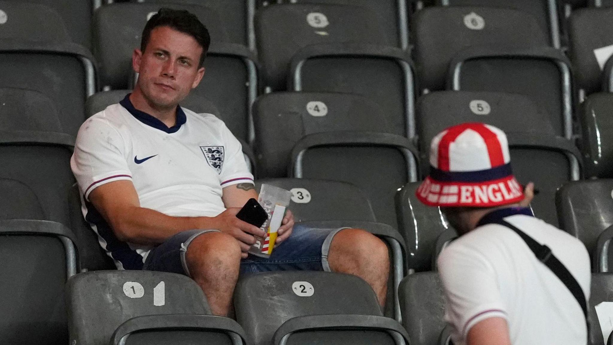 A spectator in a white England shirt and denim shorts sits alone in the stands looking dejected. Another person can be seen on a lower row to his right, wearing a white shirt and England bucket hat