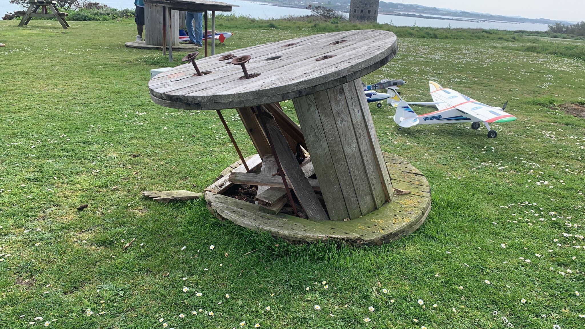 Damage to a cable drum which had been turned on its side and used as a table. The wooden structure is leaning on its side. Planks, in between the drum, have been damaged. It has been placed on a grass field. Model planes can be seen in the background.
