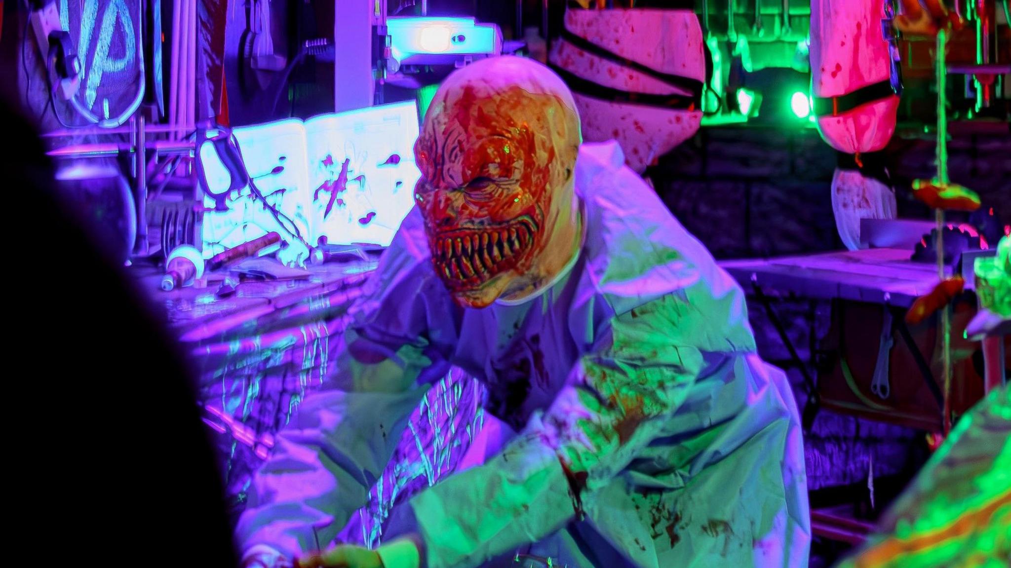 A man with a ghoulish bloody face mask with pointed teeth and wearing bloody scrubs frightens a crowd. 