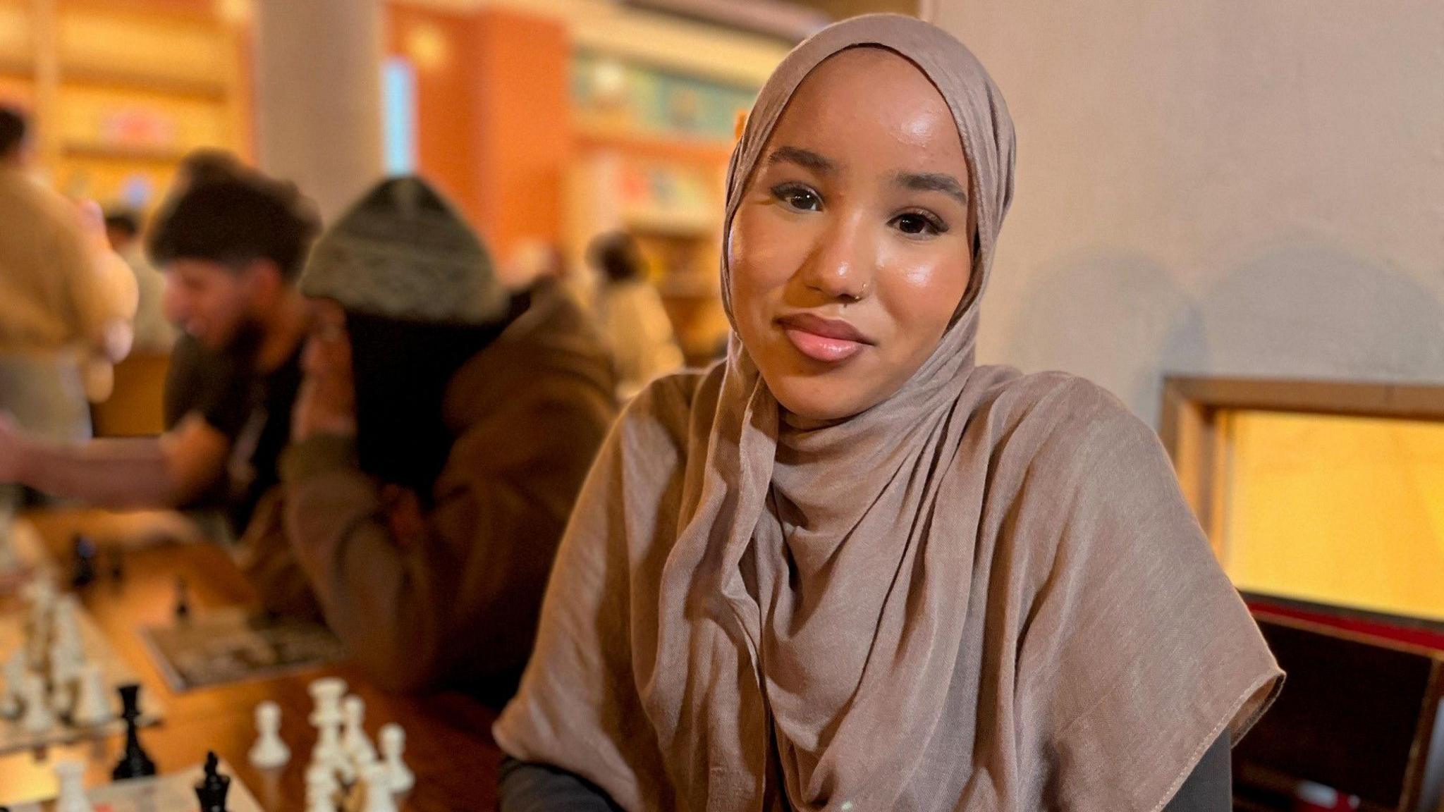 A young woman wearing a light brown hijab smiles into the camera. On the table in front of her are chess boards and two or three more young people are staring intently down at their boards.