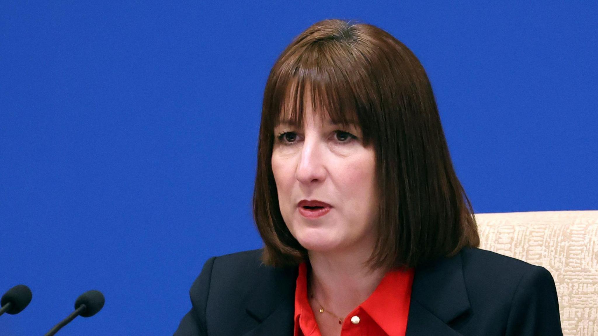 Rachel Reeves sitting at a press conference , with microphones in front of her. She has brown hair, and is wearing a dark jacket with red top. The background wall is blue. 