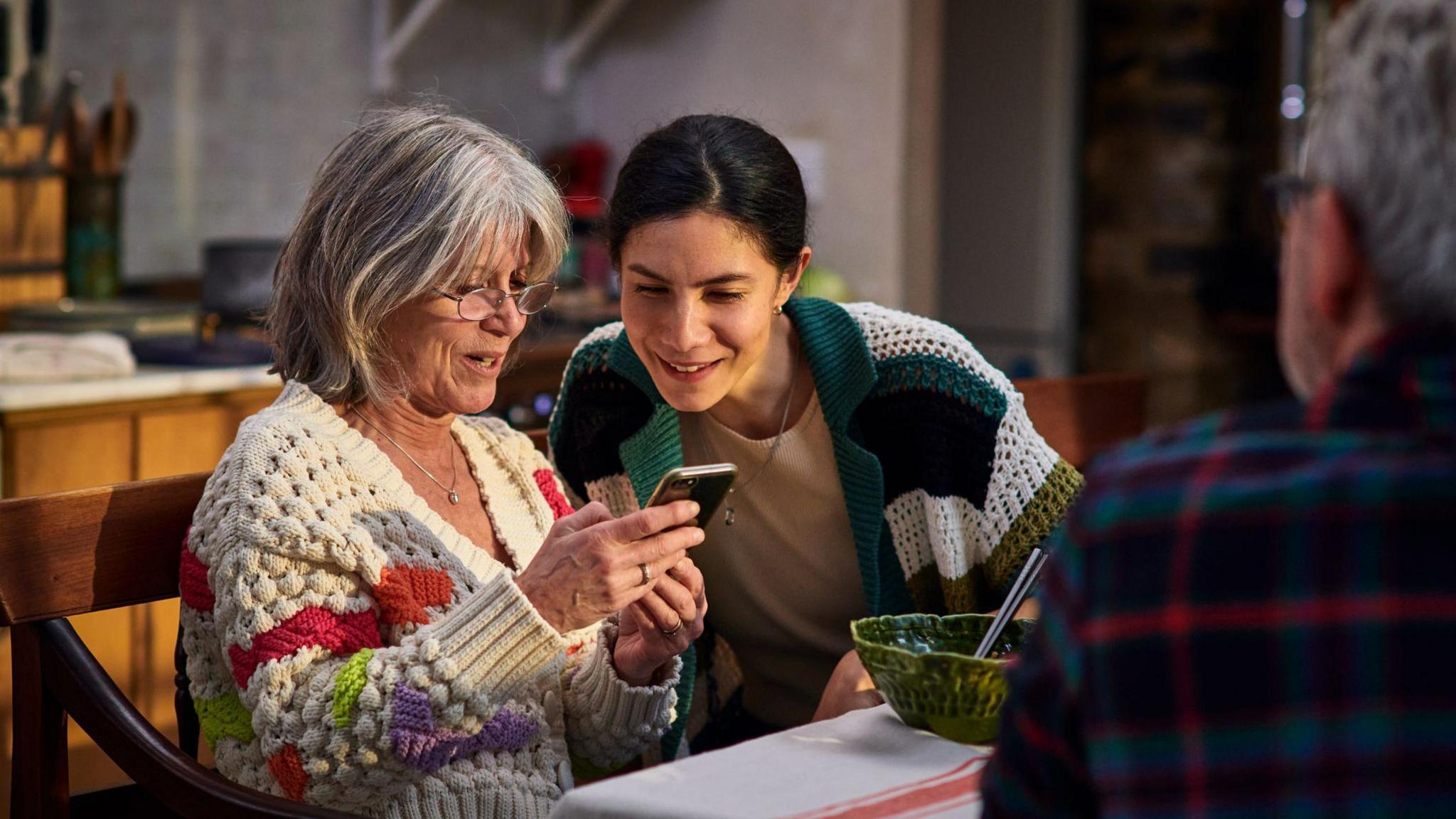 Mother and adult daughter sit at a table looking at a phone, with an older man in the foreground and a bowl on the table.